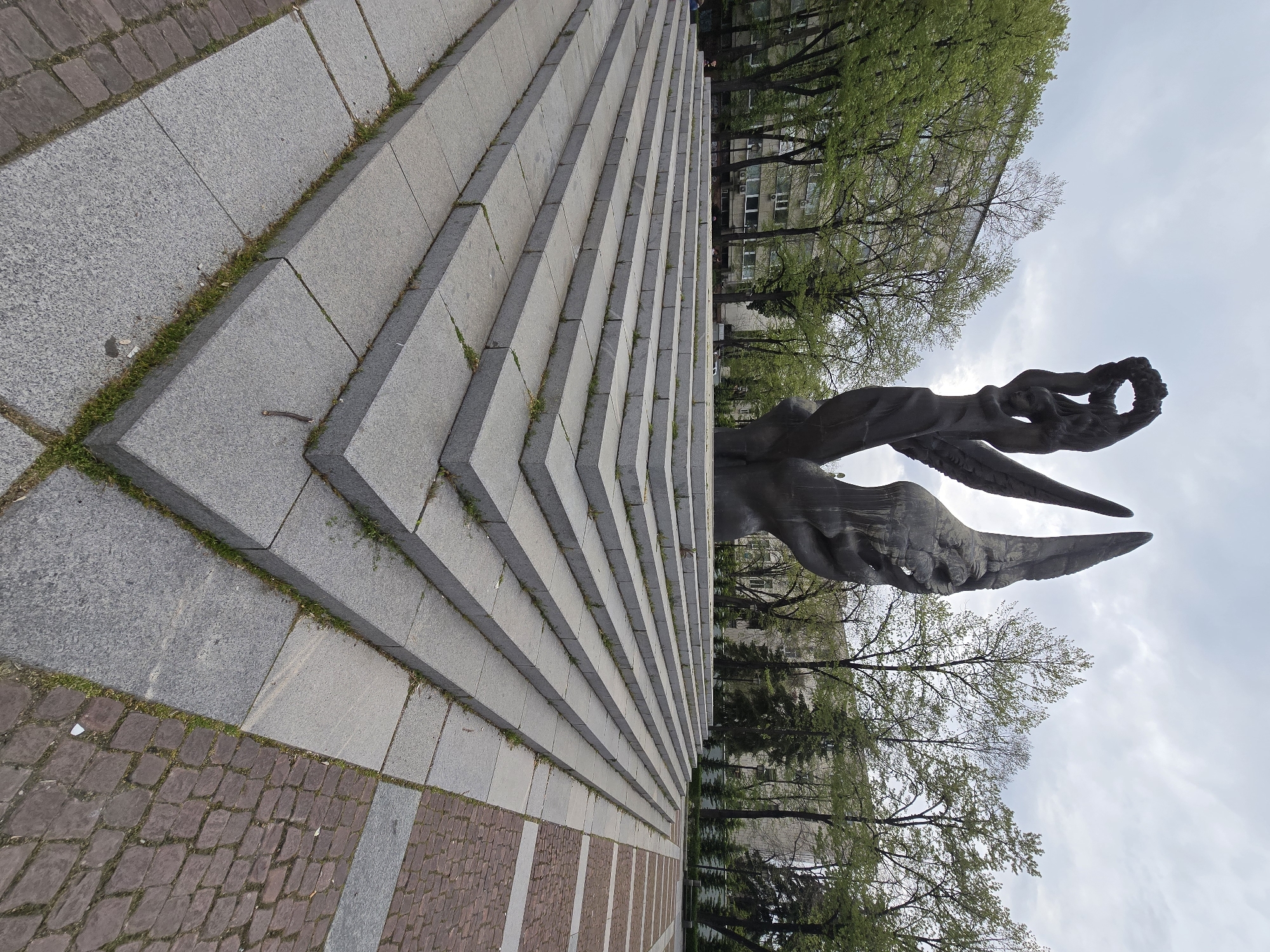 The image shows a low-angle, ground-level view of a monument or sculpture. The monument features a dark, possibly bronze or stone, figure with wings and a human-like form holding a wreath. The sculpture is positioned to the right of the frame.
To the left are a series of gray stone steps, or possibly a retaining wall, creating a sharp diagonal line. The steps are constructed of individual stone blocks with grass growing in some of the seams. The bottom of the steps is lined with a brick or cobblestone pavement in reddish-brown hues, interspersed with gray stone strips.
In the background, behind the monument, are trees in varying shades of green, suggesting foliage typical of springtime. There is a building partially visible behind the trees with windows and a facade suggesting an urban setting.
The sky is overcast and cloudy, with light filtering through the clouds. The location is listed as Unknown, Unknown. No people are visible in the image.