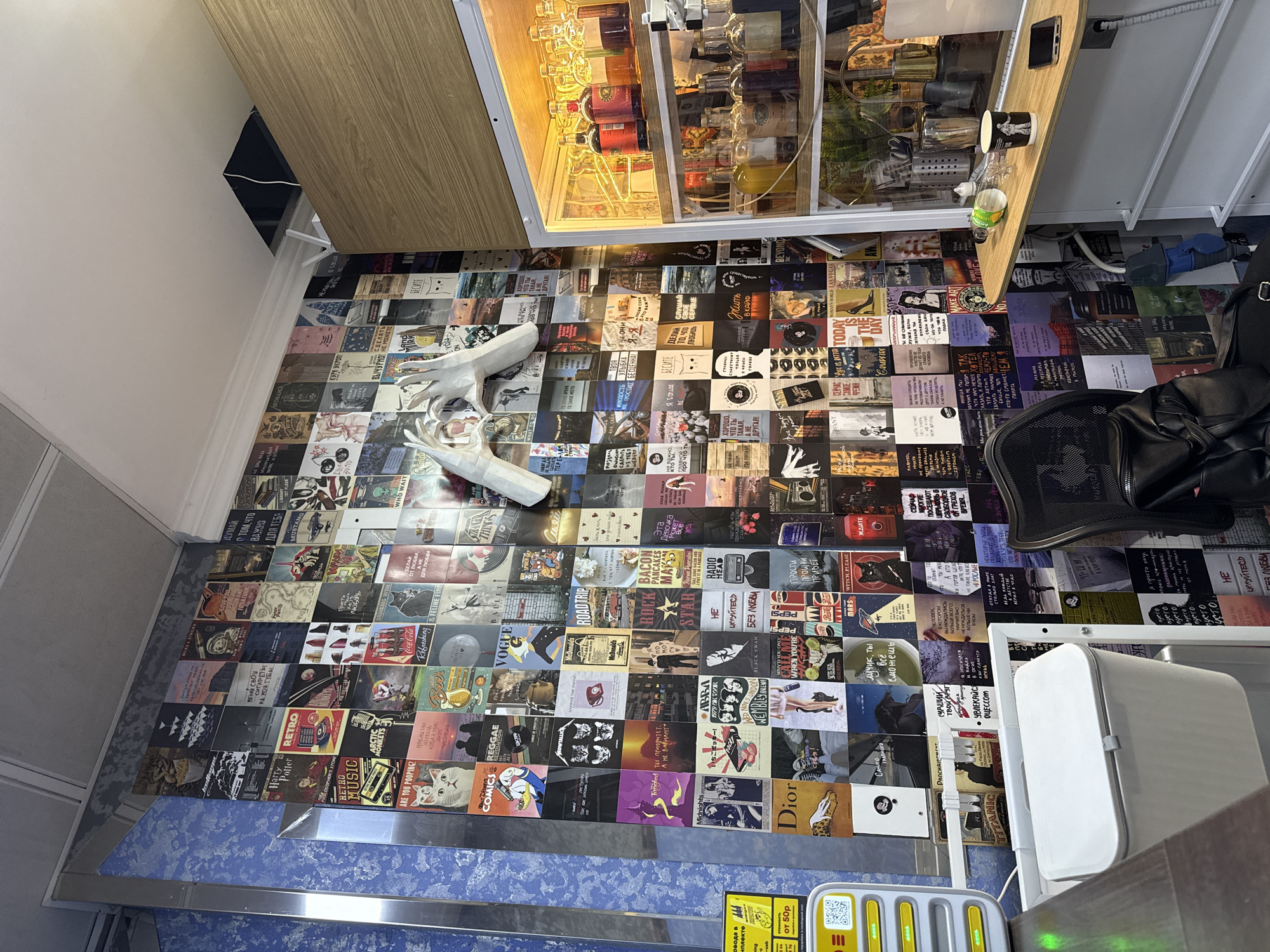 Captured from a high-angle perspective, the scene presents an interior space with a distinctive aesthetic. The floor is covered with a patchwork of album covers and posters, serving as a base for various objects.
A white deer sculpture lies on the floor, adding a touch of artistic flair. In the upper area, there is a display cabinet filled with bottles and glasses, possibly a bar setup. A black leather chair sits to the right, suggesting a seating area.
Near the bottom, there is a blue-marbled surface with some ticketing machines. The room's walls appear to be light-colored. The location could potentially be a commercial or creative space in Moscow, Russia, given the style and the Russian text on the floor coverings.