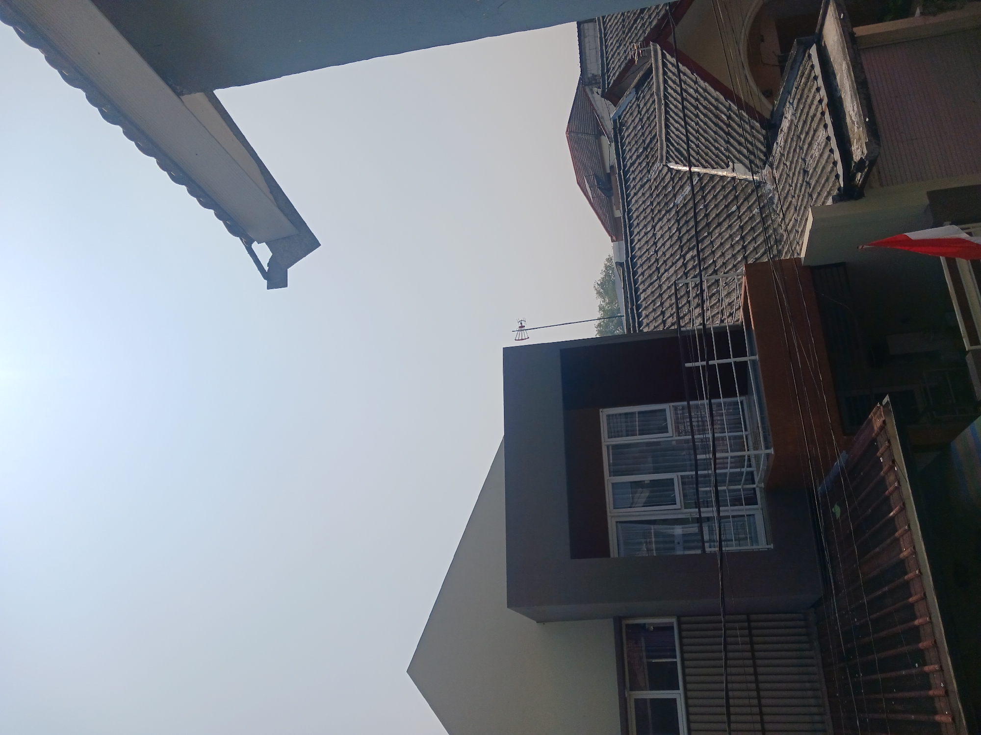 The image captures a view of rooftops and buildings in Watugede, Indonesia, under a clear, hazy sky. The foreground features a portion of a dark grey wall and a section of a tiled roof. Further back, there are multiple tiled roofs of varying conditions, some appearing older and weathered, others newer. A modern building with a dark brown rectangular protrusion containing large windows and a white railing is visible. To the left, another building with a light beige angled roof and horizontal slatted siding is seen, with windows covered by curtains. An antenna is mounted on a thin pole above the tiled roofs. In the mid-ground, parts of other residential buildings with red-tiled roofs are present. A small section of the Indonesian flag, red and white, is visible in the lower right portion of the image. The overall scene suggests a densely populated residential area.