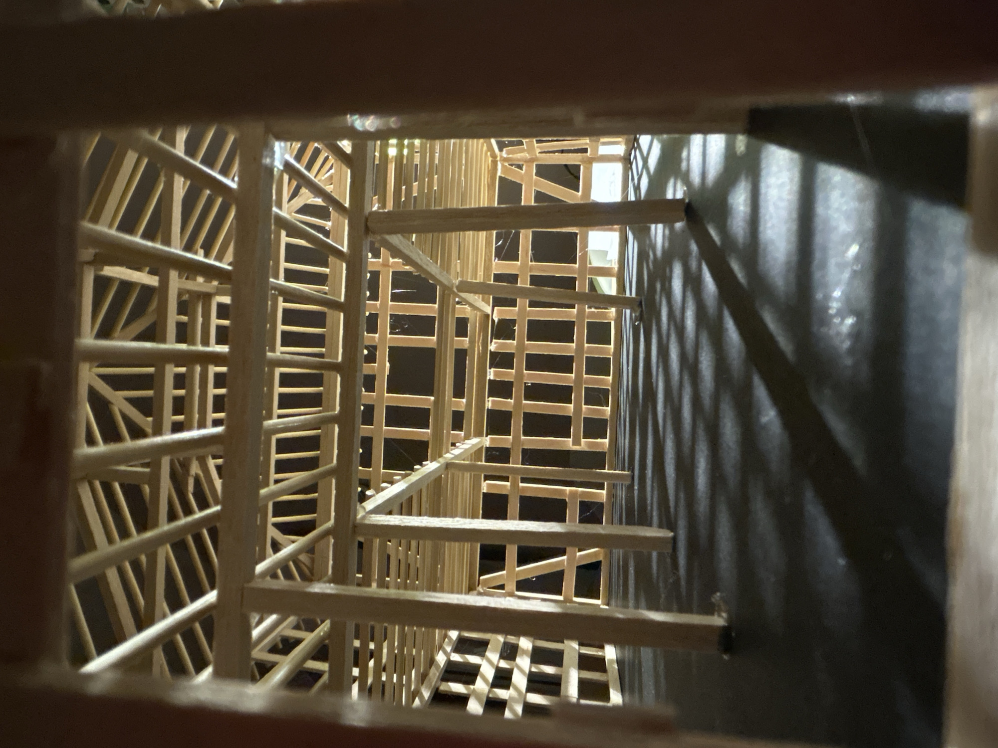 This is an indoor, eye-level, close-up shot of a model structure made of thin wooden sticks. The model appears to be a multi-level building with a complex internal framework, resembling a house or barn under construction. The lighting creates distinct shadows on the dark surface below the structure, suggesting a light source from above and to the left. The image is taken from within the structure, looking outwards through the wooden beams and supports. There is no visible indication of the city of Sofia, Bulgaria, in this image.