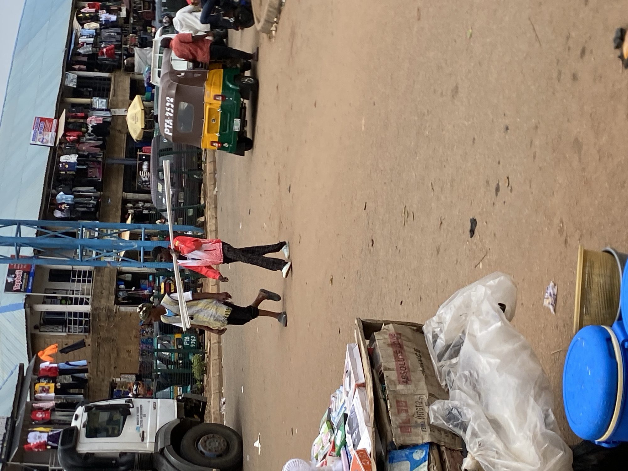This image captures a vibrant street scene in Bukuru, Nigeria. The foreground is dominated by a white truck and a collection of goods, including a large blue plastic container and a box labeled "ABSOLUTE LOVE" and "STERING: 6157GT-2." The middle ground features two men walking on a dusty road. One man, wearing a yellow and white patterned garment and a cap, carries what appears to be metal rods. The other man, in a pink shirt and dark pants, walks ahead.
Behind the men, several auto-rickshaws, also known as "keke napep," are parked or moving. One prominently displays "PTA-7532" and another "PTA 9363." The background reveals a bustling marketplace with shops displaying clothing and other merchandise. Colorful fabrics hang from stalls, and umbrellas are visible, suggesting a commercial area. The overall atmosphere appears to be one of lively commerce. The lighting suggests it is daytime, possibly mid-morning or afternoon, with the sun casting some shadows. The dusty road and the style of the vehicles and shops provide strong cues for the Nigerian location.