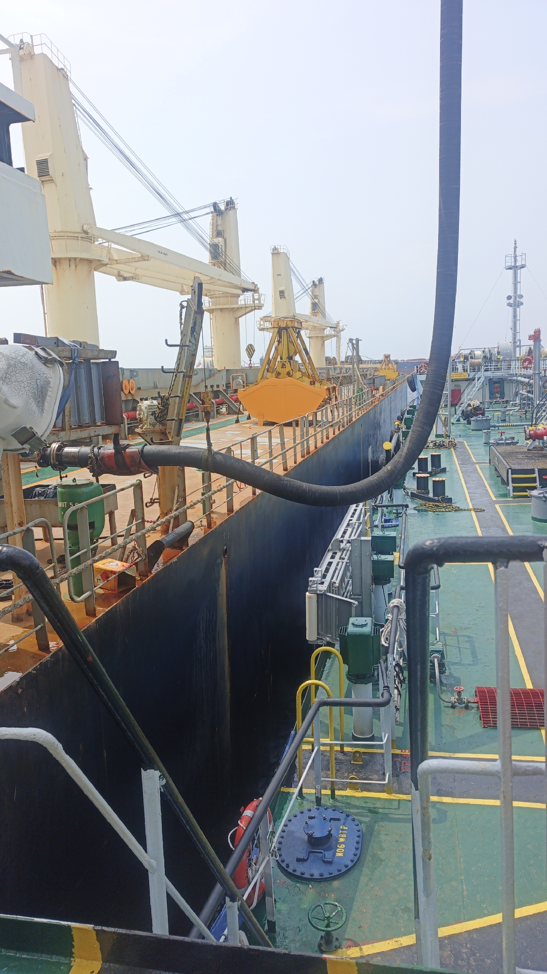 An overhead shot captures a view of two ships docked side-by-side in the Port of Singapore, Singapore, during daylight. The ship on the left, a cargo vessel, has a deep, dark hull and is laden with cargo. Cranes, some yellow and some cream-colored, are visible along its deck, with rigging and cables crisscrossing the sky. A large, thick black hose snakes from the ship on the right to the cargo ship on the left, suggesting a transfer of materials is in progress. The ship on the right appears to be a tanker, with green decking marked with yellow lines. Various pipes, valves, and equipment are scattered across its deck, including a blue circular hatch labeled "MDG WBTP." A red life preserver hangs on the side of the cargo ship. The sky is overcast, and the overall scene depicts an active industrial port environment.
