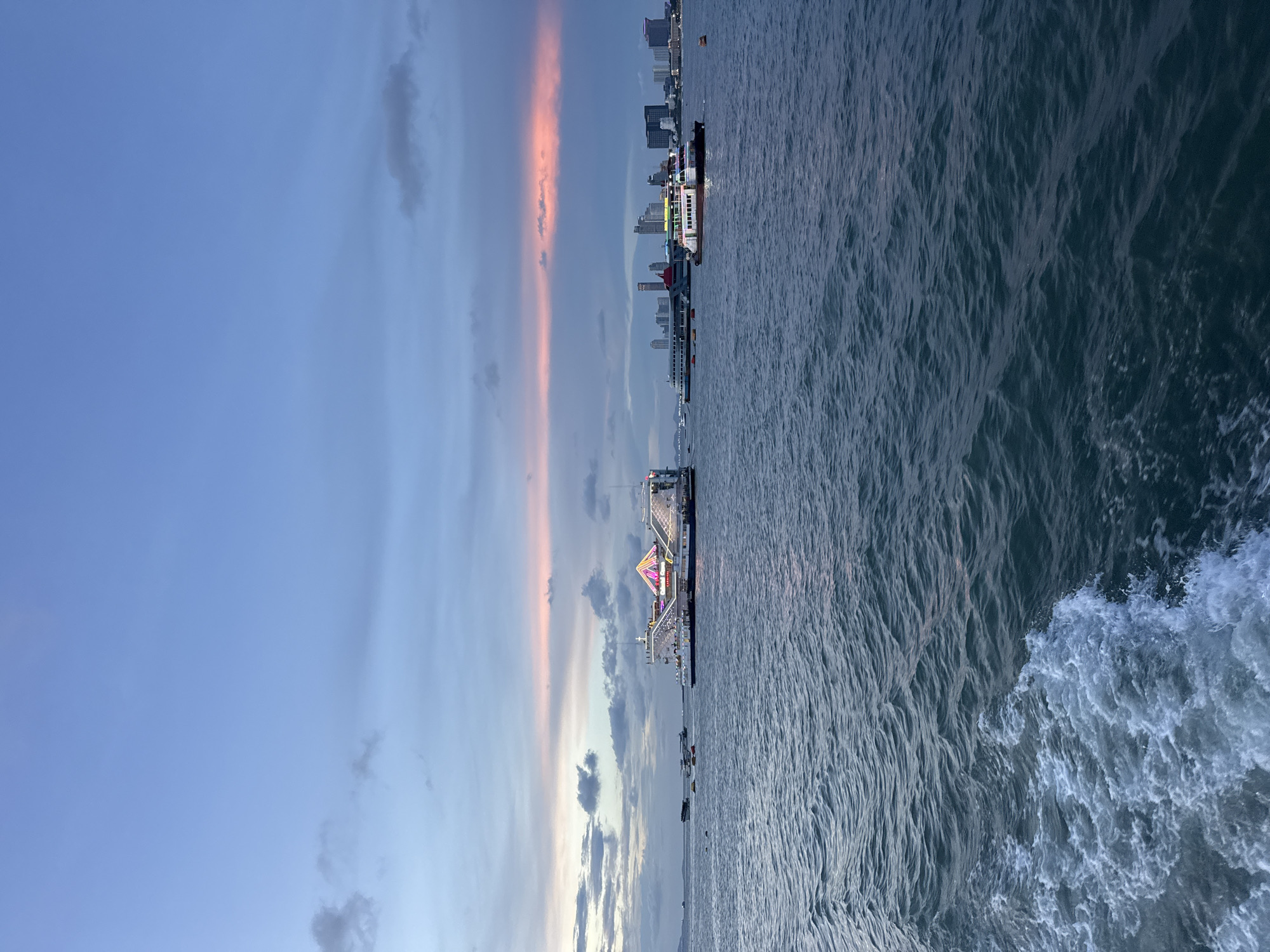 The image depicts a sunset scene on the water in Pattaya City, Thailand. The sky is a gradient of blue to pink, with distinct vertical streaks of pink clouds. On the water, there are several piers and boats. The buildings of the city are visible on the horizon to the right. The water is dark blue, showing wave patterns.