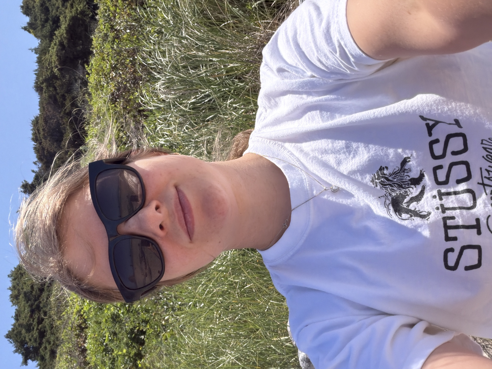 A young person with blond hair, wearing black sunglasses and a white t-shirt with a black lion graphic and the word "STUSSY" in black lettering, is taking a selfie outdoors. The background consists of tall, green grasses and shrubs, with a clear blue sky above. The person appears to be on a beach, as suggested by the presence of sand in the reflection of their sunglasses. The overall scene is bright and sunny, indicating it's daytime. The location is Cannon Beach, Oregon, a coastal city in the United States, known for its natural beauty and beaches.