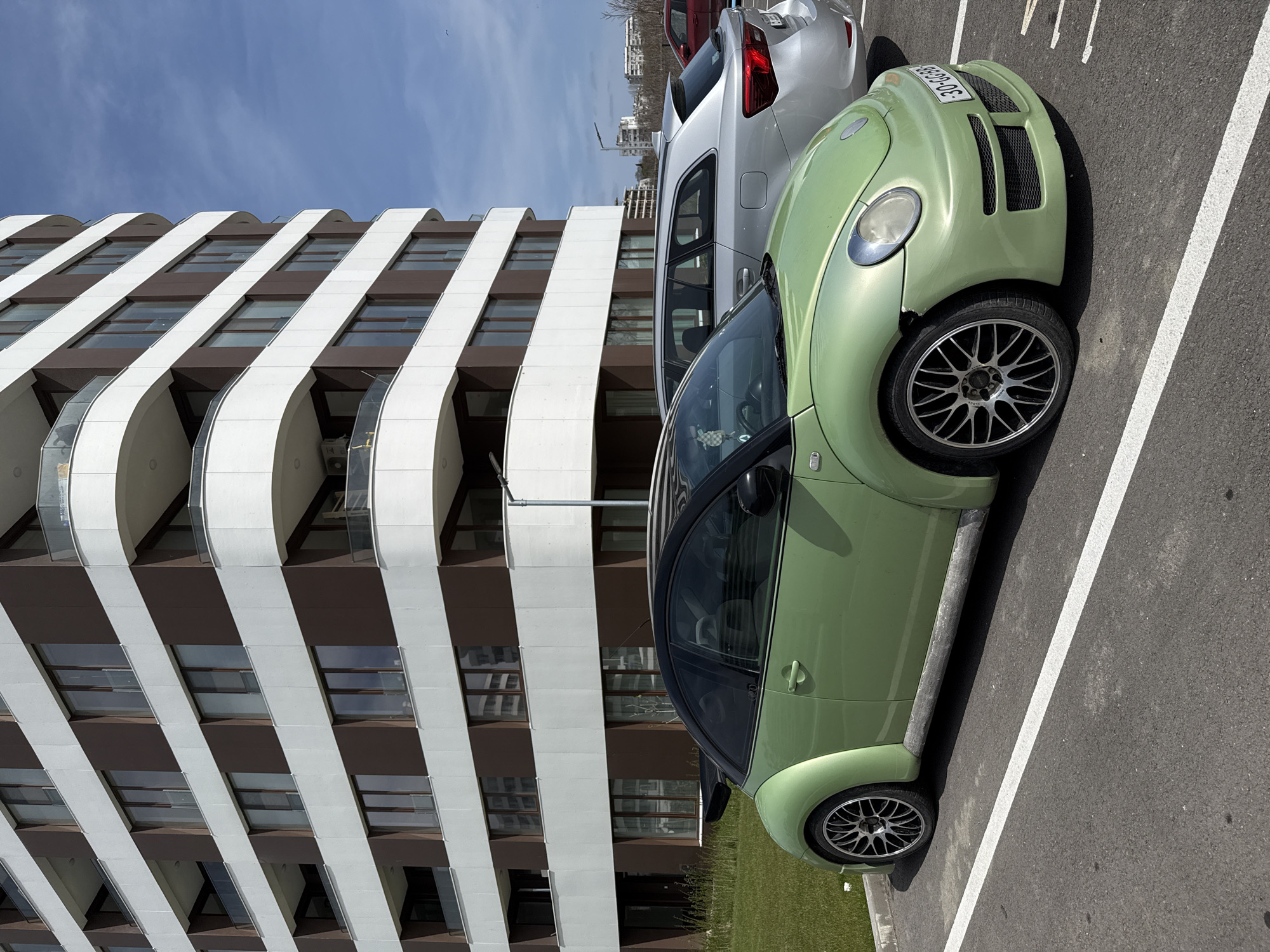 The image shows a green Volkswagen Beetle parked in a parking space next to a silver SUV. The building in the background has horizontal stripes of white and brown, with curved balconies. The sky is blue. The location is likely in Varna, Bulgaria, based on the license plate of the green car, which indicates the Varna region. The scene appears to be a typical parking area outside of a building, with cars parked in designated spaces. There are no visible people in the image.