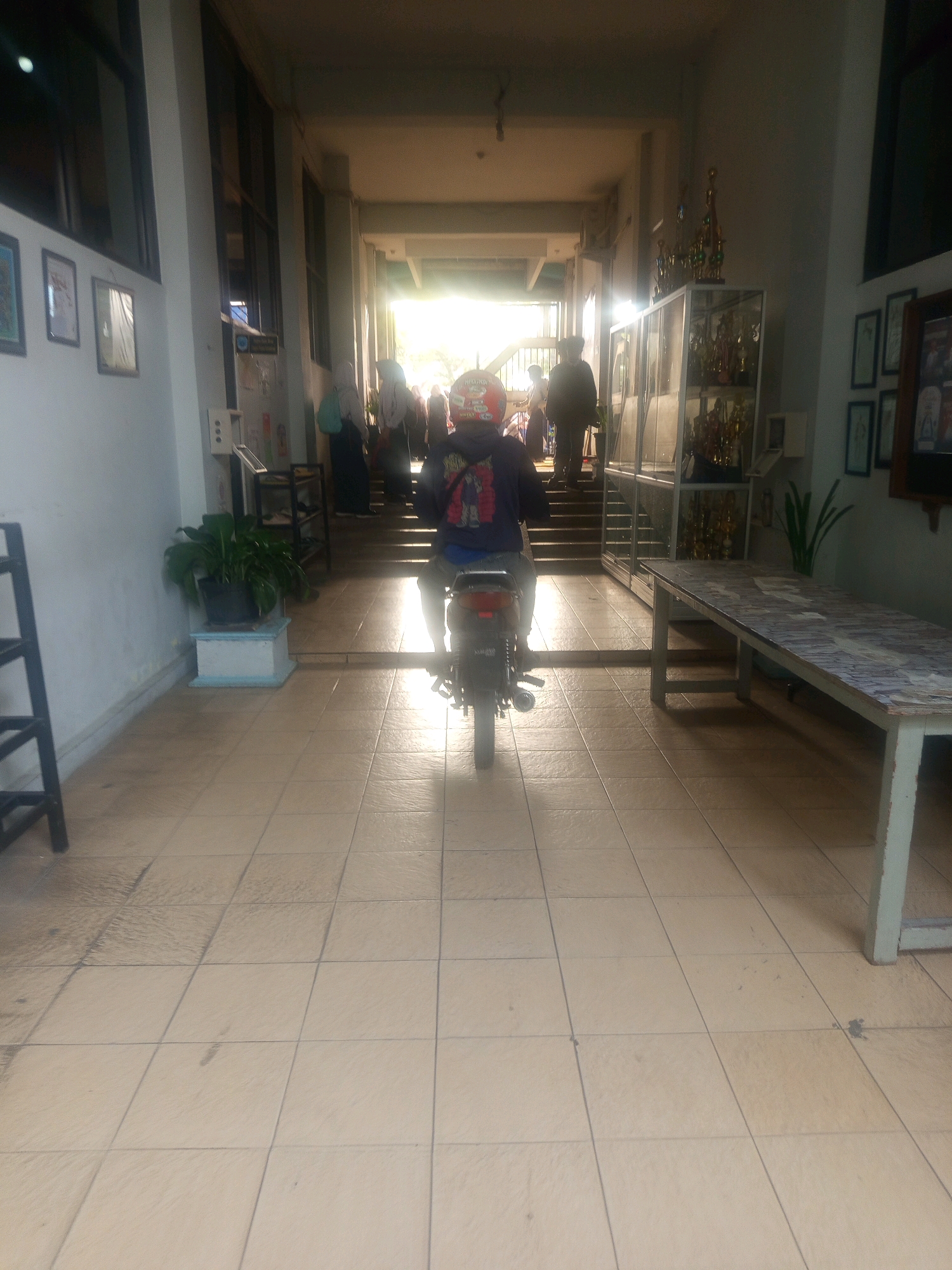 A person rides a motorcycle down a tiled hallway toward a brightly lit exit. The hallway has columns on the left and a glass display case filled with trophies on the right. Several framed pictures hang on the wall to the left of the hallway. A potted plant sits near the entrance to the hallway. The hallway appears to be inside a building, possibly a school or university, in Malang, Indonesia.