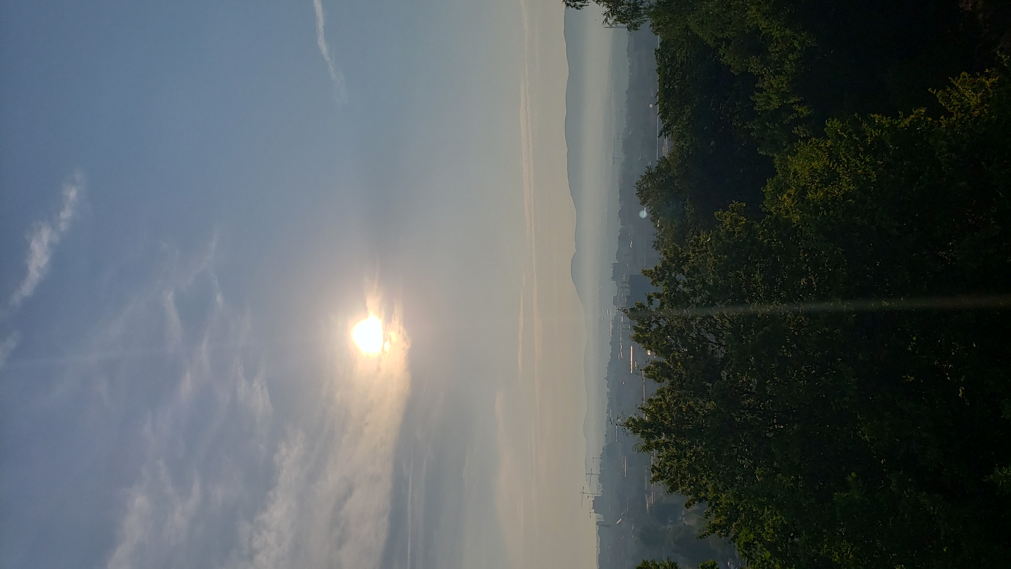 The image captures a panoramic view of the city of Prešov, Slovakia, during what appears to be early morning or late afternoon, given the low angle of the sun and the hazy atmosphere. The sun, a bright, diffused orb, is partially obscured by thin clouds in the upper left quadrant of the sky. The sky transitions from a pale blue at the top to a lighter, almost white hue near the horizon.
Below the sky, a hazy band of mist or low clouds stretches across the landscape, softening the distant features. A city skyline is visible through this haze, characterized by numerous buildings and construction cranes, suggesting ongoing development. In the far distance, a range of gently sloping mountains forms the backdrop, also softened by the atmospheric conditions.
In the foreground, the upper canopy of lush green trees dominates the lower portion of the image. These trees, with their dense foliage, create a natural frame for the cityscape. A faint vertical lens flare extends downwards from the sun, creating a subtle light streak in the sky and through the foliage. No people or specific actions are discernible in the scene. The overall impression is one of a serene, elevated view over a city at the edge of a natural landscape, enveloped in soft light and atmospheric haze.