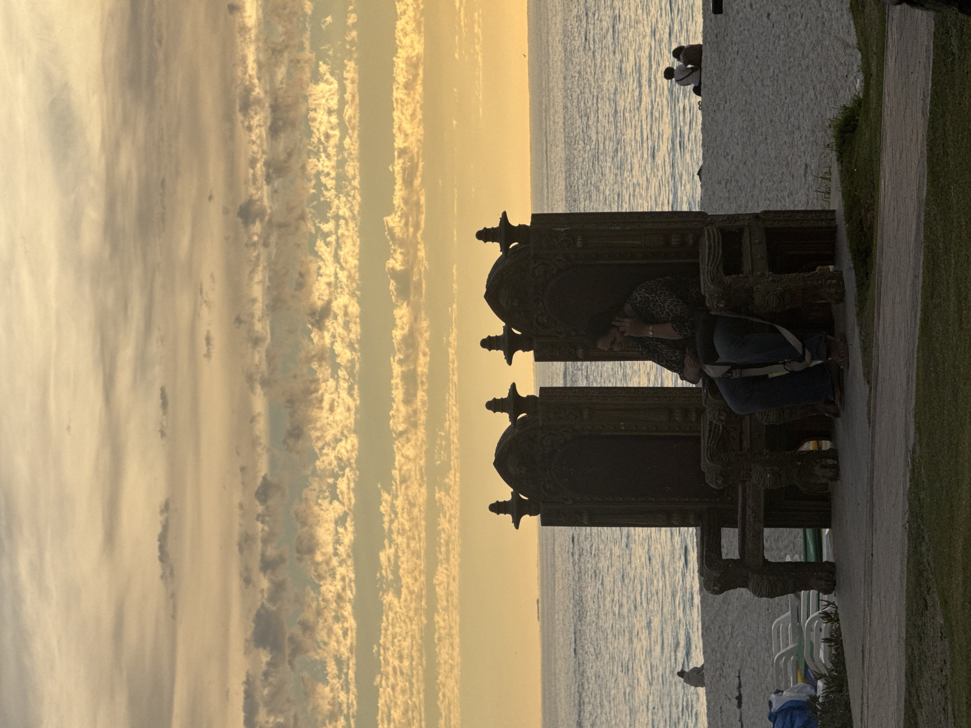 A medium shot captures a silhouetted scene at sunset on the beach near the waterfront in Cape Town, South Africa. A woman wearing jeans and a dark print jacket reclines in one of a pair of dark, ornate throne-like chairs placed at the edge of the shoreline. She leans her head on her hand, seeming pensive. The ocean is directly behind the chair, and the sky is a vivid blend of yellow, orange, and gold. A line of clouds runs parallel to the water, adding texture to the sky. In the far distance, several people sit on the beach. The scene is serene, casting a golden hue over the area.