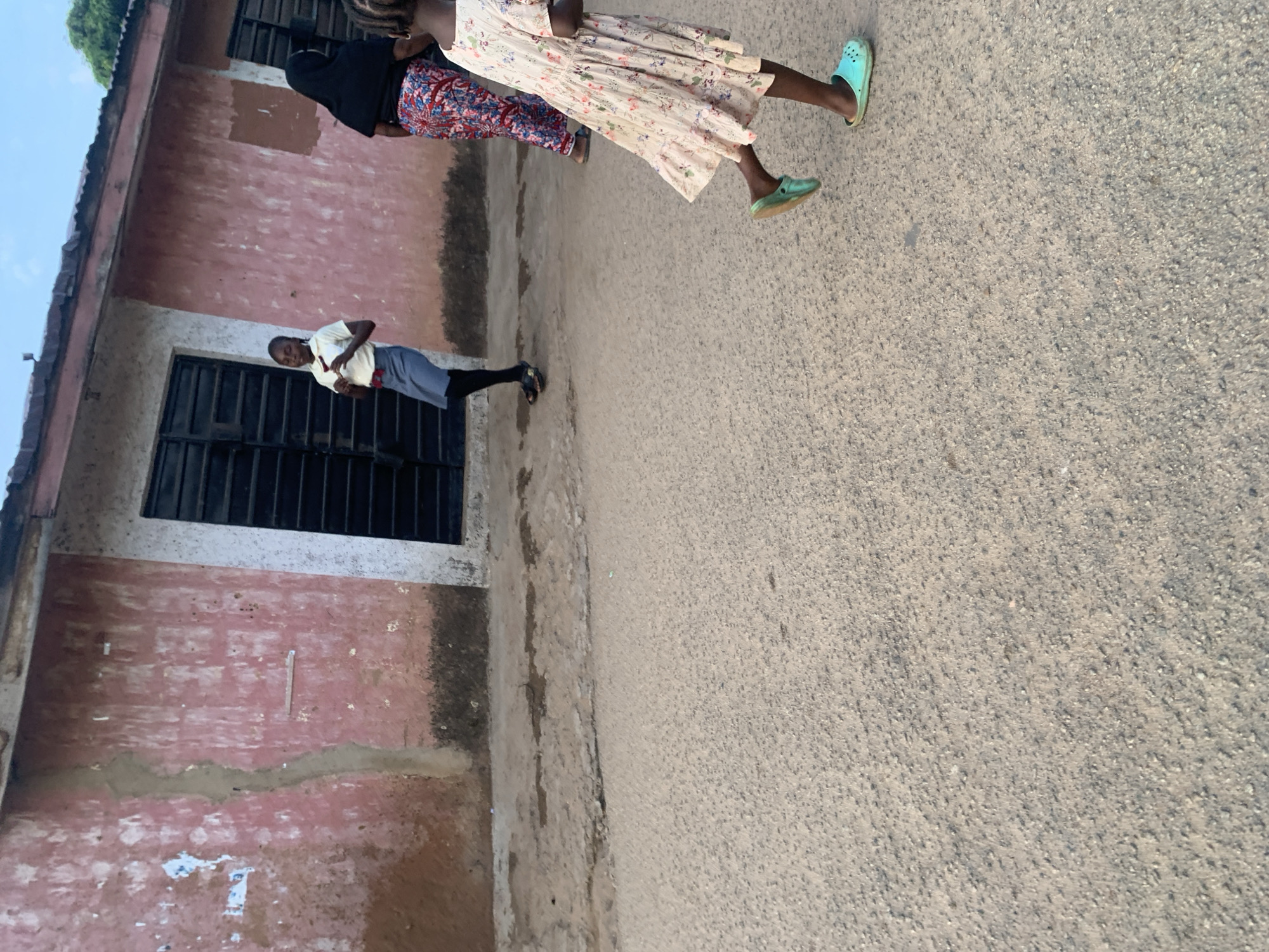 The image depicts a street scene in Jos, Nigeria. A young girl in a light-colored, floral-print dress and turquoise clogs is in the foreground, running to the right, her body angled away from the camera. Behind her, another young girl in a white shirt and gray skirt with black leggings and dark sandals is standing, facing left and holding something in her hand. Further to the right, a woman in a black hijab and a colorful patterned skirt walks away from the camera. The background features a weathered, pink building with dark window coverings and a narrow sidewalk. The street surface is asphalt with visible gravel. The sky above is a pale blue.
