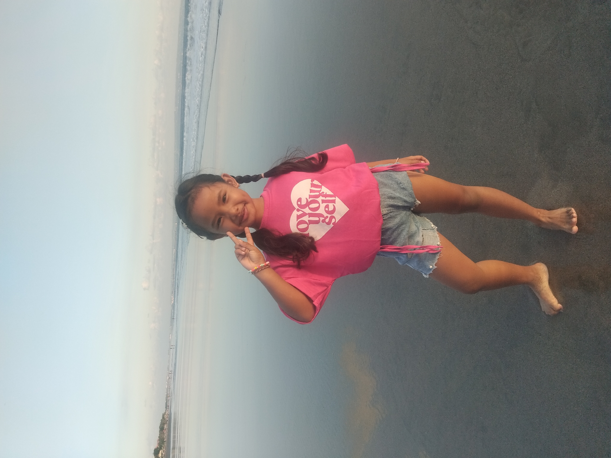 An image, presented in a portrait orientation but depicting a scene that appears to be horizontally composed, features a young girl lying on a sandy beach. The girl, with dark hair styled in two long braids, is wearing a bright pink t-shirt and light blue denim shorts. On her t-shirt, a white heart graphic displays the text 'Love Your Self'. She also has colorful beaded bracelets on her left wrist.
She is positioned on her side, facing the viewer with a broad smile, and holds her left hand up to make a peace sign. Her bare feet are visible, resting on the dark, wet sand or shoreline.
The background shows the ocean with gentle waves breaking on the shore. The sky is a soft, light blue, transitioning to a lighter hue at the horizon, where faint land features or distant structures can be seen. The lighting suggests either late afternoon or early morning, giving the scene a soft, diffused quality. The girl's joyful expression contributes to an overall cheerful and serene atmosphere.
