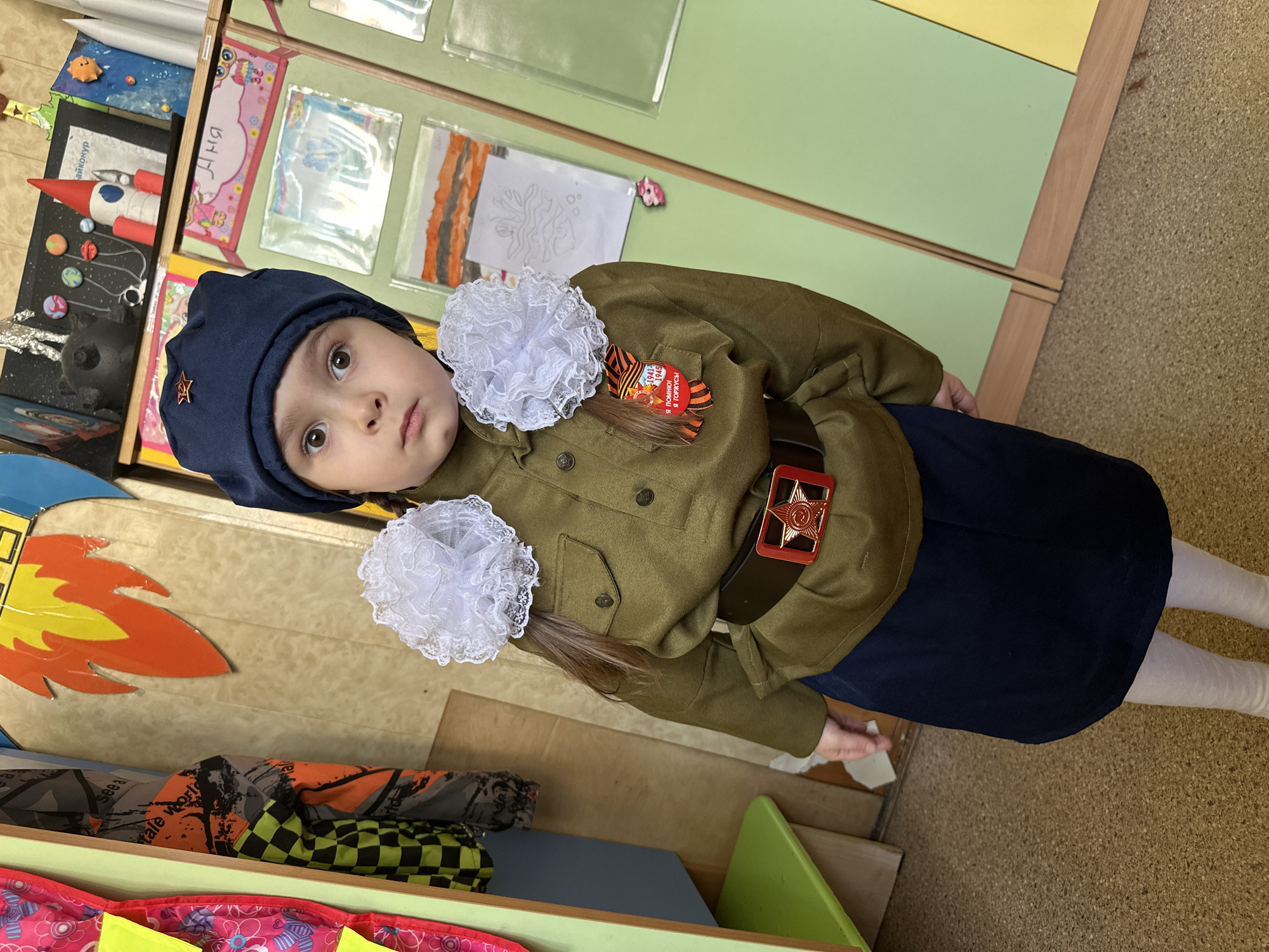 A young girl in an olive-green military uniform, a navy blue hat adorned with a red star, and white tights is standing indoors. She has white lace bows in her hair. A brown belt with a red star buckle is around her waist. A "Victory Day" ribbon is pinned to her chest. The setting includes a storage unit with children's artwork and toys. The location is Krasnogorsk, Russia.