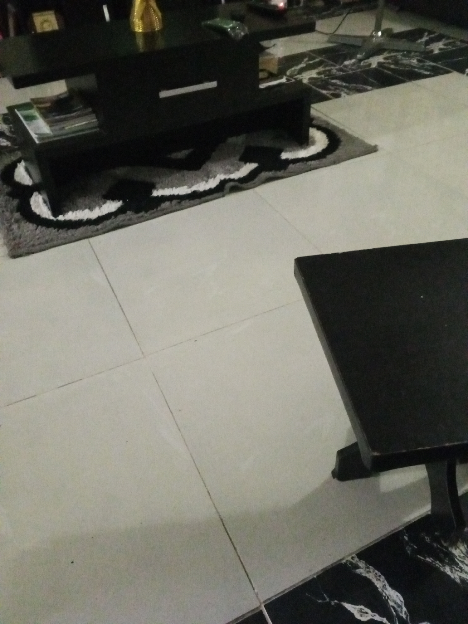 The image shows a close-up view of a modern living room interior. Two black coffee tables are visible. The table in the foreground is a simple, low-profile design with a flat top and visible legs. The second coffee table, positioned further back and to the left, is more complex, featuring a tiered design with magazines stored on a lower shelf. This table also has a sleek, minimalist drawer or compartment. A small, textured rug with a curved black and white pattern is placed partly under the second coffee table. The floor is tiled with large, light-colored square tiles, with some darker, patterned tiles visible in the background. The overall impression is of a clean, contemporary living space, likely in an urban residential setting within Howayi, Nigeria, given the context. The lighting suggests an indoor environment, possibly with artificial light or natural light from a window not visible in the frame. There are no people or active events depicted in the scene; it is a still interior shot.