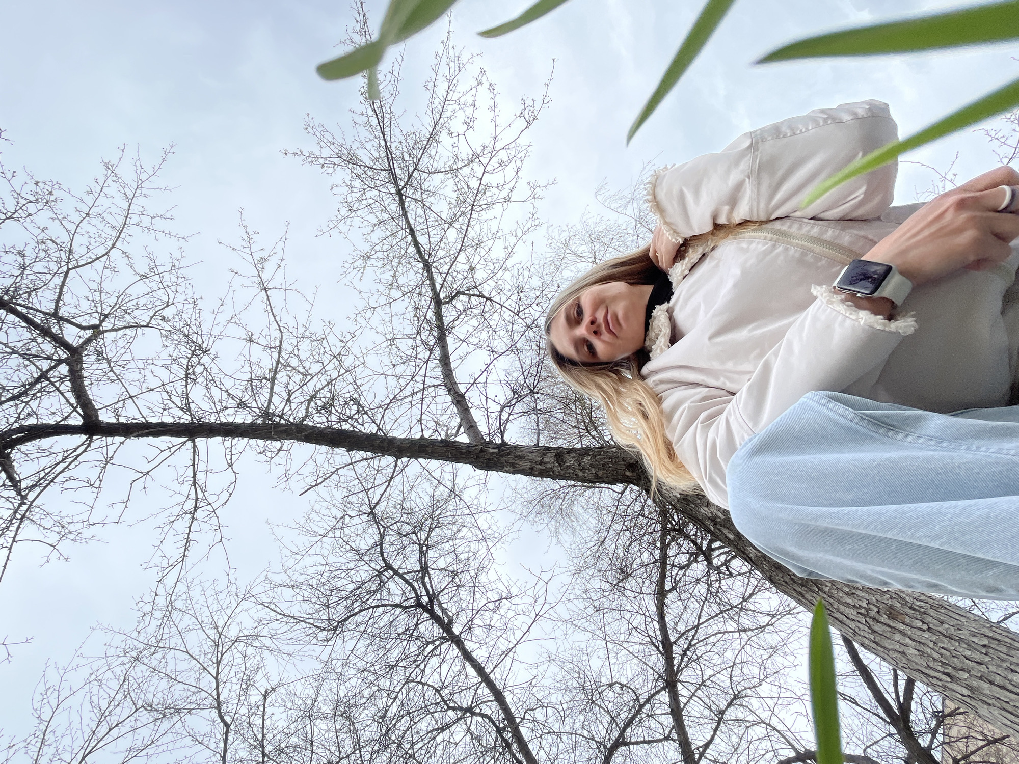 A low-angle shot captures a young woman positioned against a tree trunk, looking down towards the camera, with a backdrop of bare trees and an overcast sky. The perspective is from directly below her, giving a unique upward view.
The main subject, a woman with long blonde hair, is wearing a light-colored, possibly cream or off-white, puffer jacket with ruffled trim on the collar and cuffs, a dark top underneath, and light blue jeans. A silver-colored watch with a light band is visible on her left wrist, and she wears a ring on her right hand, which is held near her neck. Her expression is neutral as she gazes into the lens.
The setting is outdoors, likely in a park or wooded area in Chaikovsky, Russia, given the context. The numerous deciduous trees are completely devoid of leaves, suggesting late autumn, winter, or early spring. The sky is uniformly cloudy and bright, indicating daytime without direct sunlight. Some out-of-focus green leaves are visible in the very foreground, slightly obscuring the bottom and upper right portions of the frame, hinting at a new season or evergreen foliage nearby. No text is visible in the image.