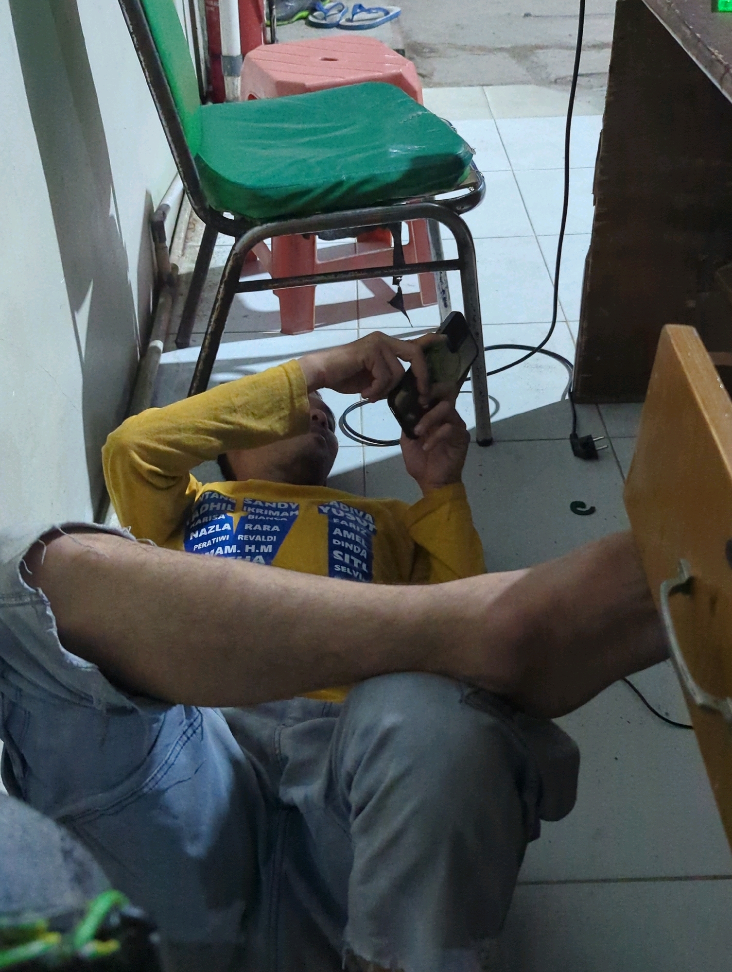 A person is lying on their back on the floor with their legs extended, facing a desk. The person is wearing a yellow long-sleeved shirt with blue lettering and light blue jeans. They are holding a dark-colored mobile phone in their right hand, looking at the screen. The person's left arm is bent at the elbow, resting behind their head. The environment appears to be indoors, with a tiled floor and a white wall. There is a green-seated chair with a metal frame next to the person, and a red plastic stool behind it. A dark-colored electrical cord is visible on the floor, plugged into an outlet near the desk. The desk is made of dark wood. The scene is illuminated by ambient lighting, casting shadows. This image was captured in Batu Belaman, Indonesia.