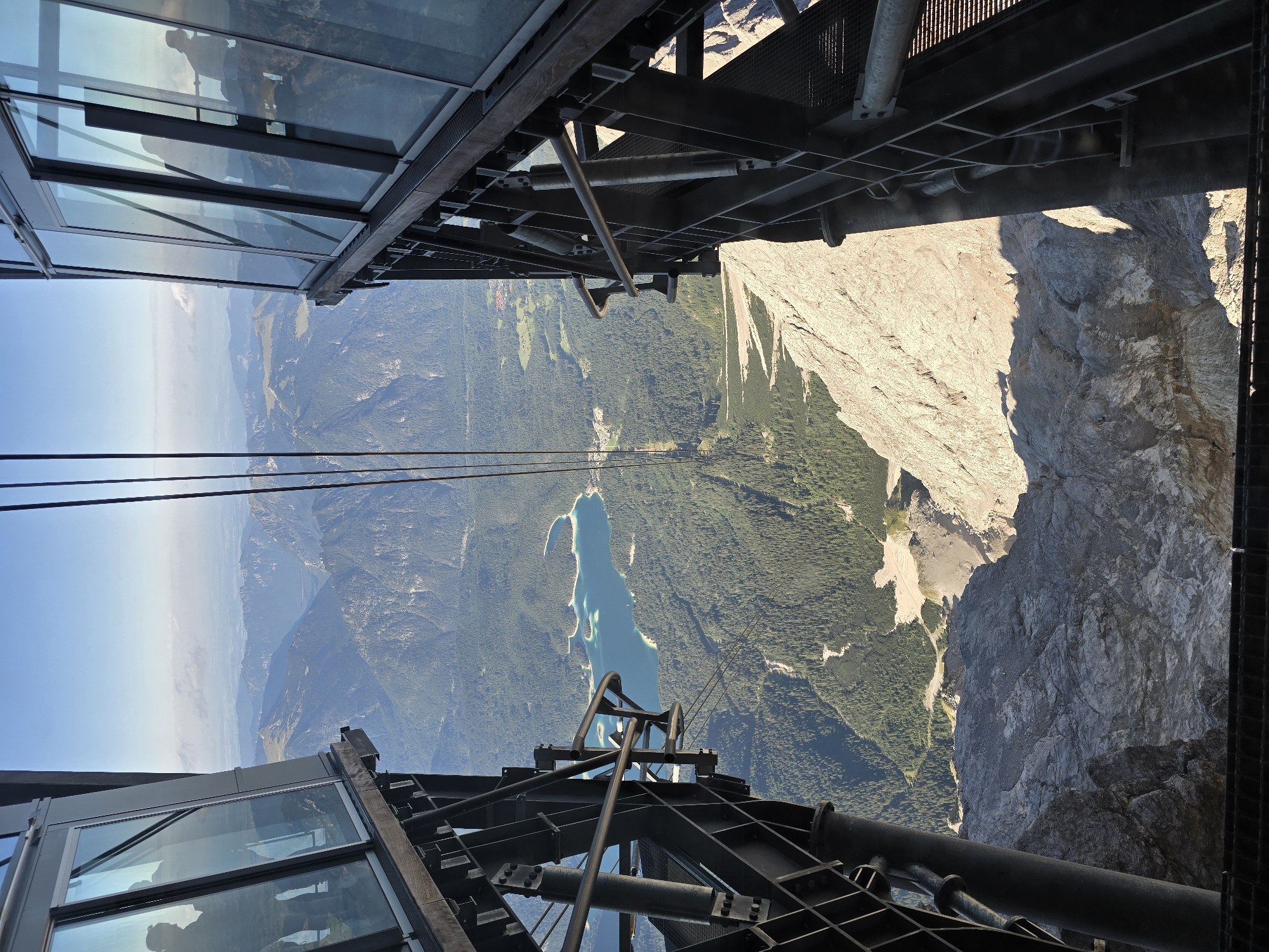 This image captures a view from a cable car ascending a mountain, likely the Zugspitze, near Garmisch-Partenkirchen, Germany. The camera is positioned inside the cable car, looking downwards and outwards. The cable car itself features large glass windows, reflecting the sky and mountain scenery. Structural metal supports and railings are visible in the foreground, framing the expansive vista.
Below, a bright turquoise lake, possibly the Eibsee, is nestled amidst a dense, green pine forest that covers the lower slopes of the surrounding mountains. The distant landscape is characterized by undulating, mist-shrouded mountain ranges under a clear blue sky. The rocky, sunlit face of a steep mountain cliff occupies the bottom portion of the image, emphasizing the significant altitude. Silhouettes of people can be seen in the reflections on the glass windows of the cable car, indicating the presence of other passengers. The lighting suggests it is a bright, sunny day, with shadows cast by the mountain peaks.