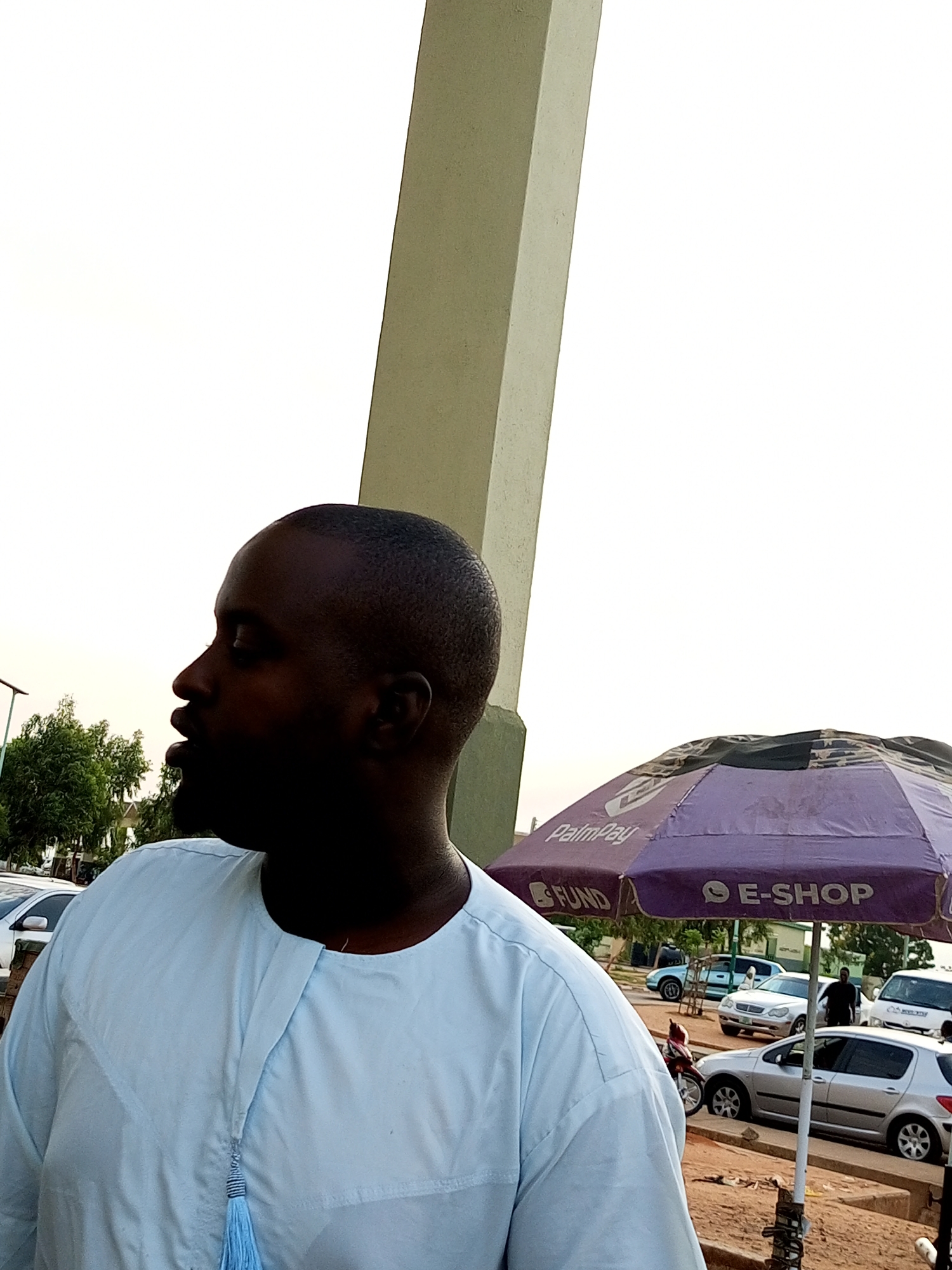 The image depicts an outdoor scene in Gombe, Nigeria, likely during the day. In the foreground, a man is shown from the chest up, wearing a light blue traditional outfit with a tassel detail. He is facing to his left, with his mouth slightly open. To the right and behind him, a purple umbrella with "PalmPay" and "E-SHOP" branding is visible, suggesting a commercial or service area. In the background, several cars are parked on a dirt or sandy road, including silver sedans and a white van. Trees are present behind the cars, and a large, light-colored vertical structure, possibly a pillar or part of a building, is prominent on the left side of the frame. The sky is overcast or hazy.