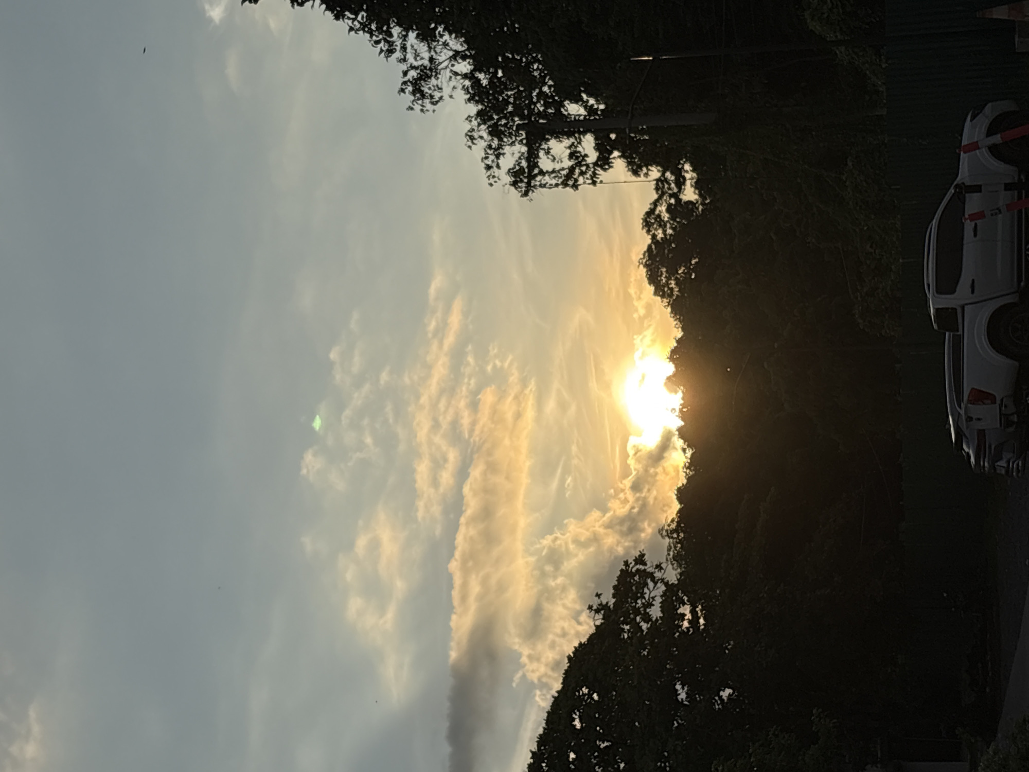 A photograph captures the sky above a densely forested area, illuminated by the setting sun. The sun, a bright orb, peeks through a gap in the clouds and tree line, casting a warm, golden glow across the sky. The clouds are a mix of white and orange, with some appearing dark and heavy, suggesting potential rain. The trees in the foreground are silhouetted against the bright sky, their dark foliage creating a stark contrast. In the lower portion of the image, two parked vehicles are visible, a white pickup truck and a darker, possibly black, SUV. The scene suggests a natural landscape within an urban or suburban setting. Based on the presence of lush tropical vegetation and the quality of light, this scene is likely located in a tropical region, such as Davao City, Philippines, known for its abundant green spaces and proximity to natural landscapes.