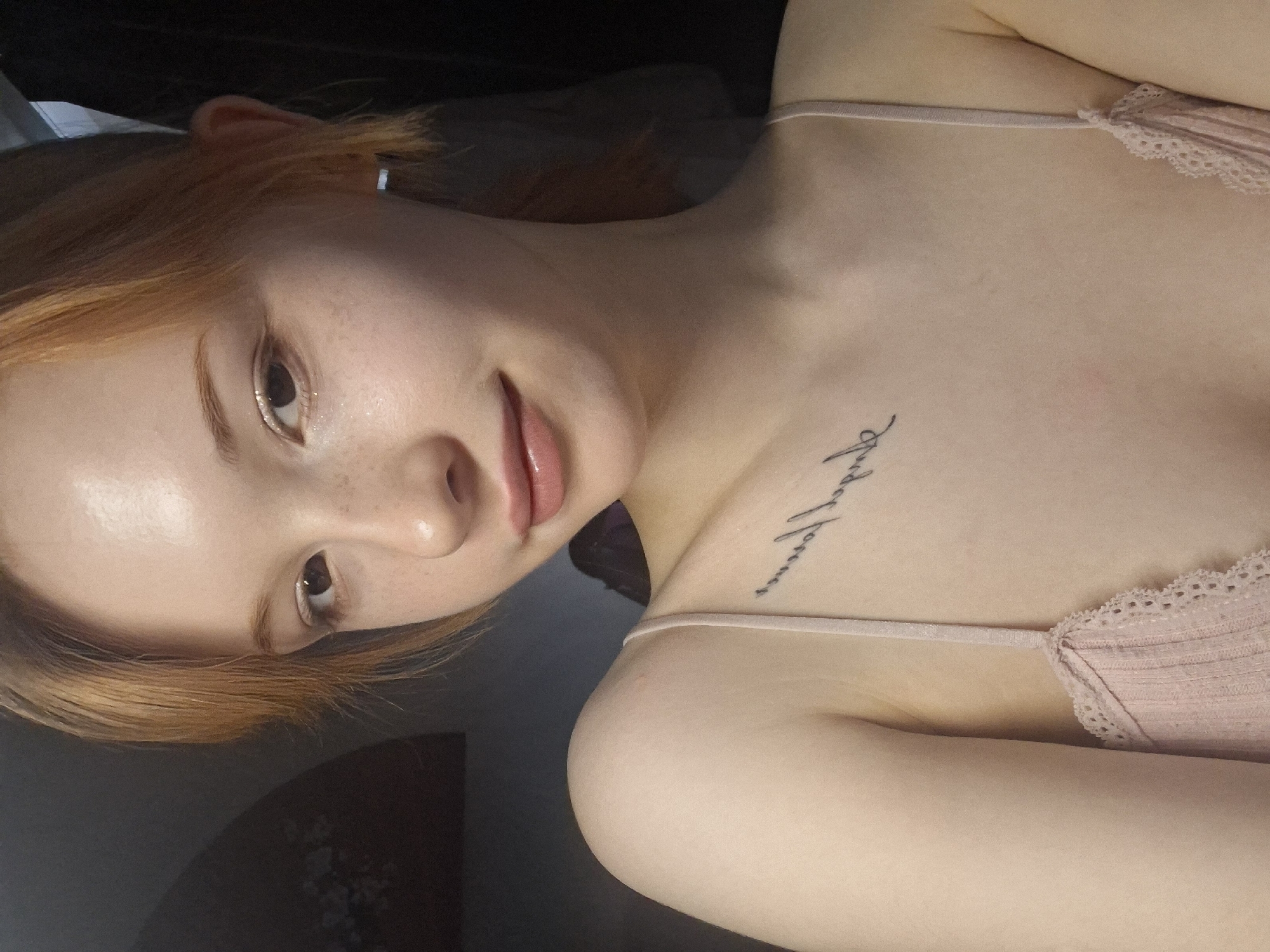 A young woman with fair skin and freckles is angled towards the camera. She has light brown/reddish hair. She's wearing a pale pink tank top with lace trim. A tattoo is visible on her upper chest/collarbone area. The background is dark. Location is not discernable from the image.