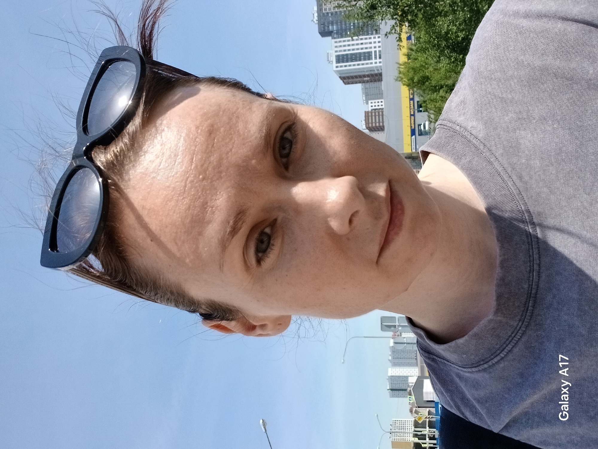 A selfie portrait shows a woman with sunglasses on her head, taken on a bright day in Yekaterinburg, Russia. Behind her are high-rise buildings and trees. The sky is blue, and there are streetlights visible. "Galaxy A17" is written on the bottom right.