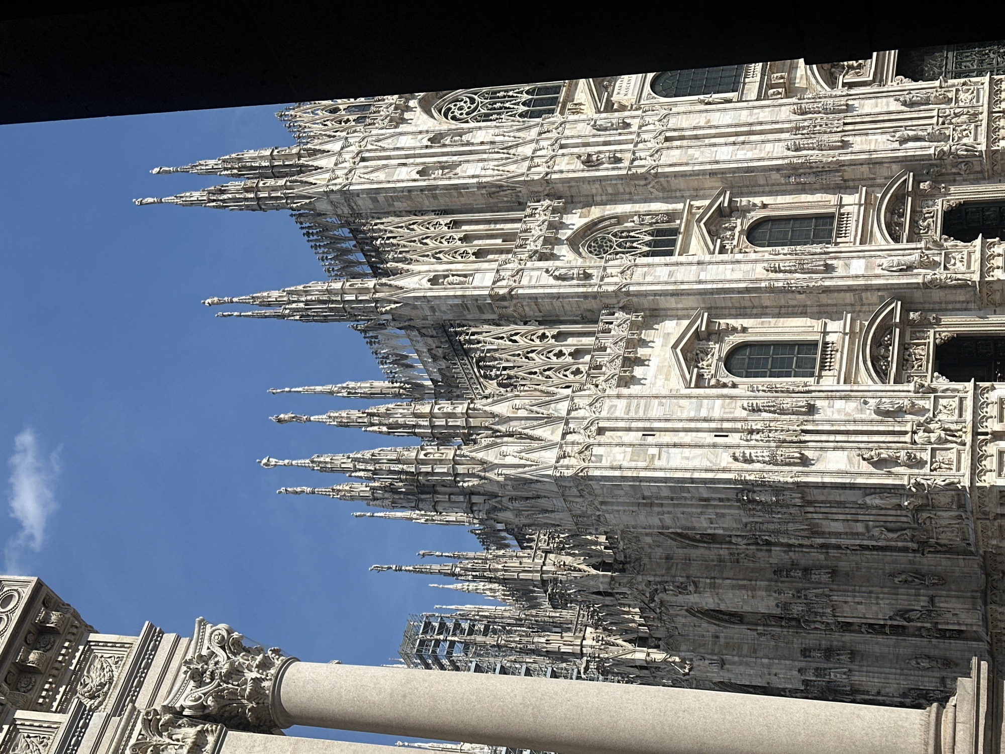The image captures a detailed, low-angle view of the Milan Cathedral (Duomo di Milano) against a clear blue sky with one small white cloud in the upper left. The cathedral's ornate Gothic architecture is prominent, featuring numerous intricately carved spires, flying buttresses, and statues crafted from light-colored marble. A section of the cathedral on the mid-left displays scaffolding, indicating ongoing maintenance. On the far left, a large, light-grey stone column with a highly decorative capital partially frames the scene, belonging to an adjacent structure. The right side of the image is framed by a dark, vertical architectural element or shadow. Bright sunlight illuminates the cathedral, casting sharp shadows that emphasize its three-dimensional details, including arched windows with elaborate tracery and sculpted figures. Text "MARIAE NASCENT" is visible on a lower section of the cathedral's facade. No individuals are present in the scene.