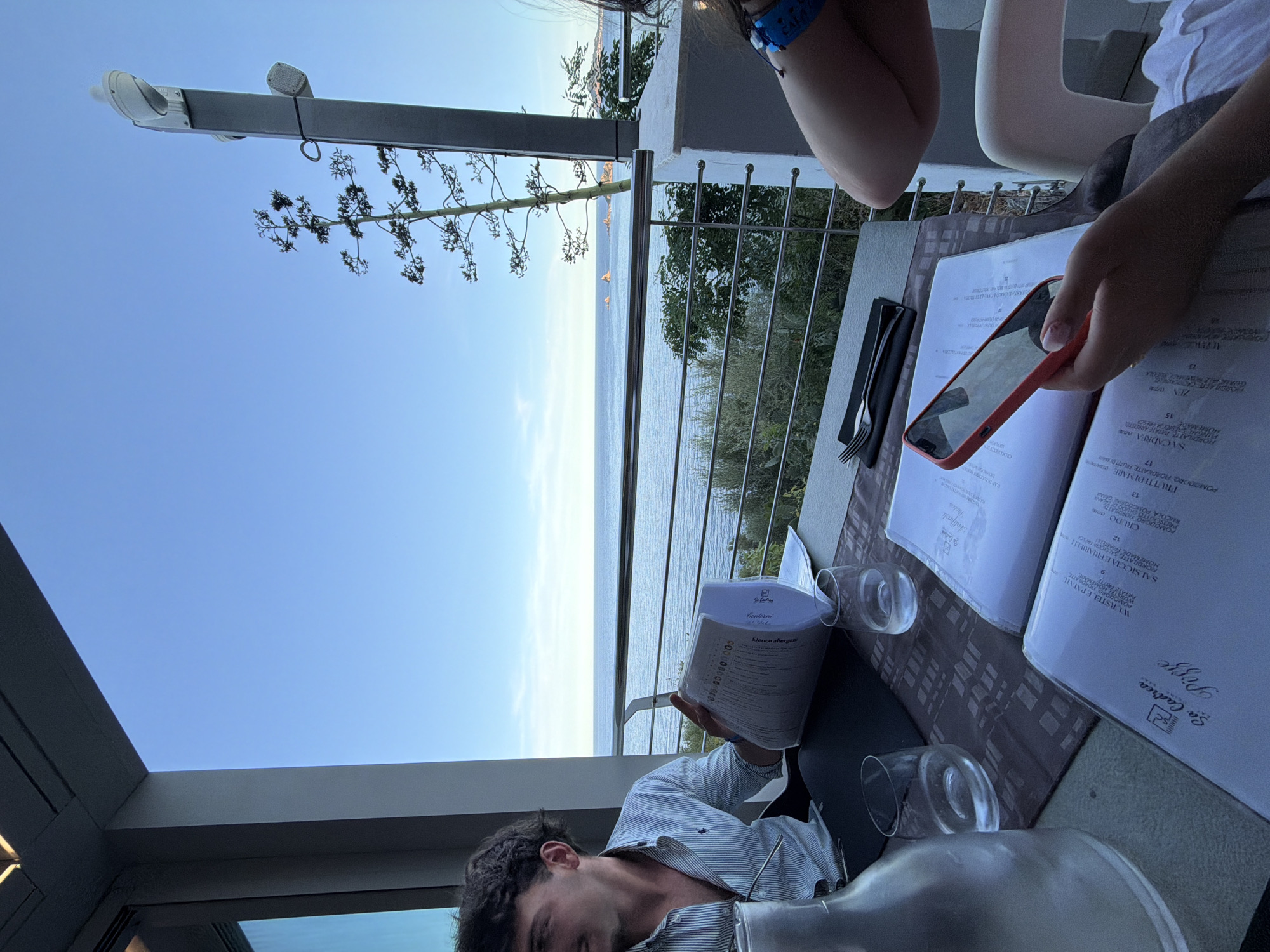 The image depicts a scene from a restaurant with a view of the sea, likely in Santa Maria Navarrese, Italy. Two individuals are seated at a table. On the left, a young man with dark, curly hair, wearing a light blue and white striped button-down shirt, is looking away from the camera. In front of him, on the table, is a menu, a glass carafe, and a partially filled glass of water. On the right side of the table, a person, whose arm and shoulder are visible, is holding a red smartphone and looking at a menu. The table also has place settings with cutlery and napkins. The restaurant appears to have an outdoor dining area with a railing overlooking the sea. The sea is calm, with a clear blue sky above and some rocky outcrops in the distance. A tall, spindly plant is visible in the background, as is a lamppost with two lights.