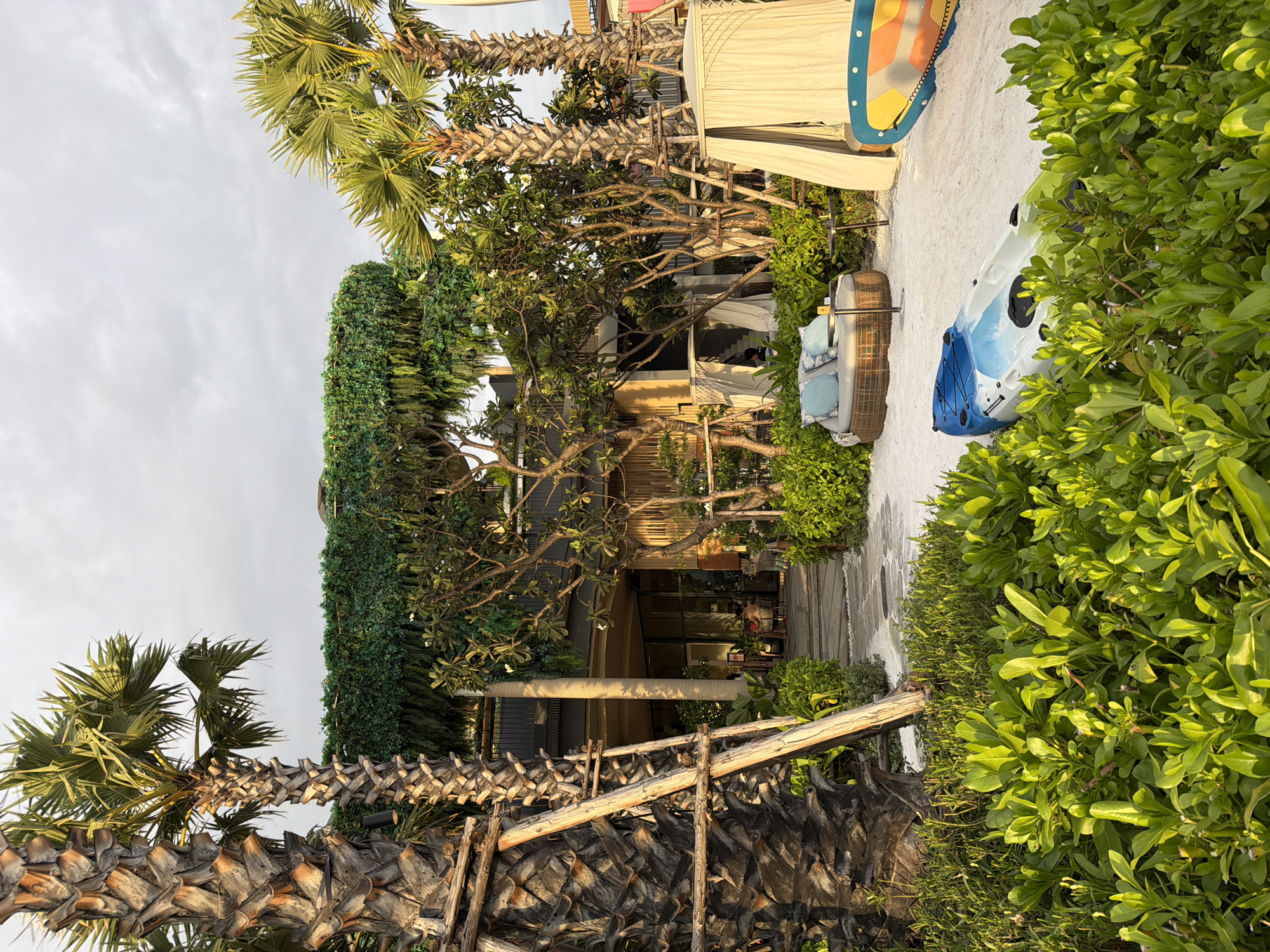 The image shows a beachfront scene in Pattaya City, Thailand. A two-story building is visible, extensively decorated with green vegetation, appearing as if the building is covered in a vertical garden. Palm trees are interspersed with the building and surrounding landscape. A sandy path leads towards the building, with greenery on either side. A blue and white kayak and a yellow and blue paddleboard are partially visible to the right of the path, suggesting the area is near a beach. The sky overhead is overcast. Some seated people are visible in the interior through large windows.