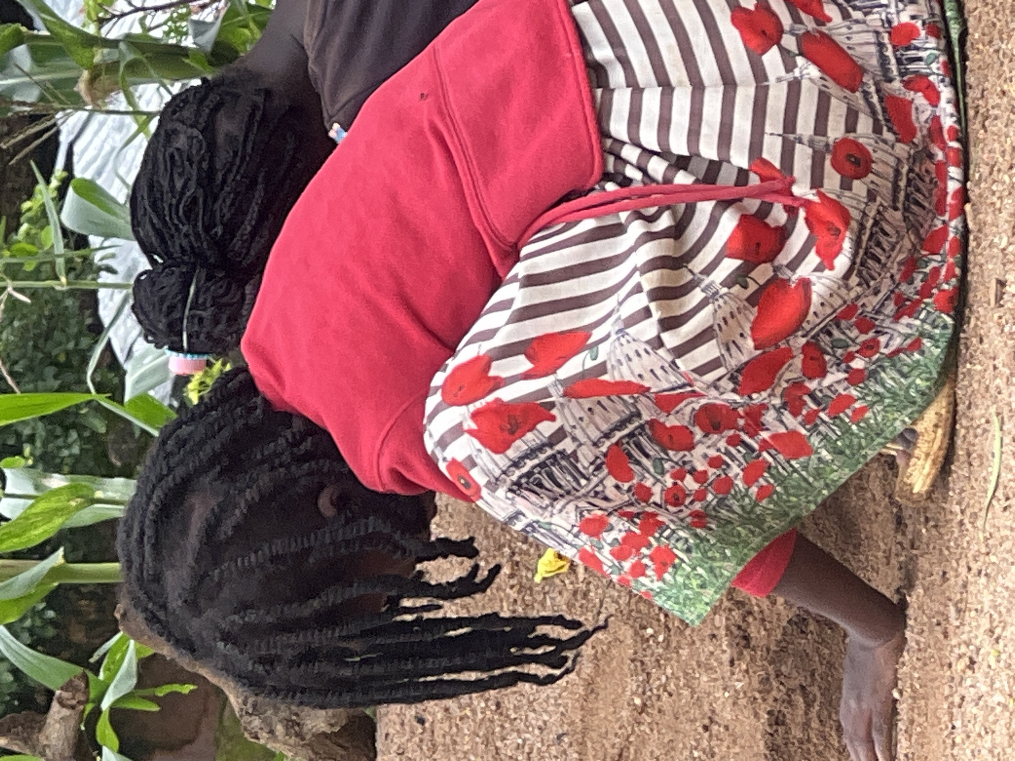 The image shows two individuals outdoors, in what appears to be a rural or semi-rural setting. The foreground features the back of a person wearing a bright red hooded sweatshirt and a skirt with a striking pattern of red poppies and a green border, over a striped design. The person is crouched down, with their left arm extended and hand resting on the sandy ground. Their hair is styled in dark braids that fall down their back.
To the right and slightly behind the first person, another individual is visible, also with dark hair styled in braids, pulled back. This second person is leaning forward, with their head resting on or near the shoulder of the first person.
The background consists of greenery, including tall stalks of what appears to be corn, and foliage. A rough, textured surface, possibly a rock or tree trunk, is visible to the left. The ground is composed of sand and small pebbles. The overall impression is of a casual outdoor scene.
While the image depicts people in an outdoor environment, there is no specific visual information within the image that allows for the identification of the location as Jos, Nigeria. The scene is consistent with many rural or peri-urban environments.