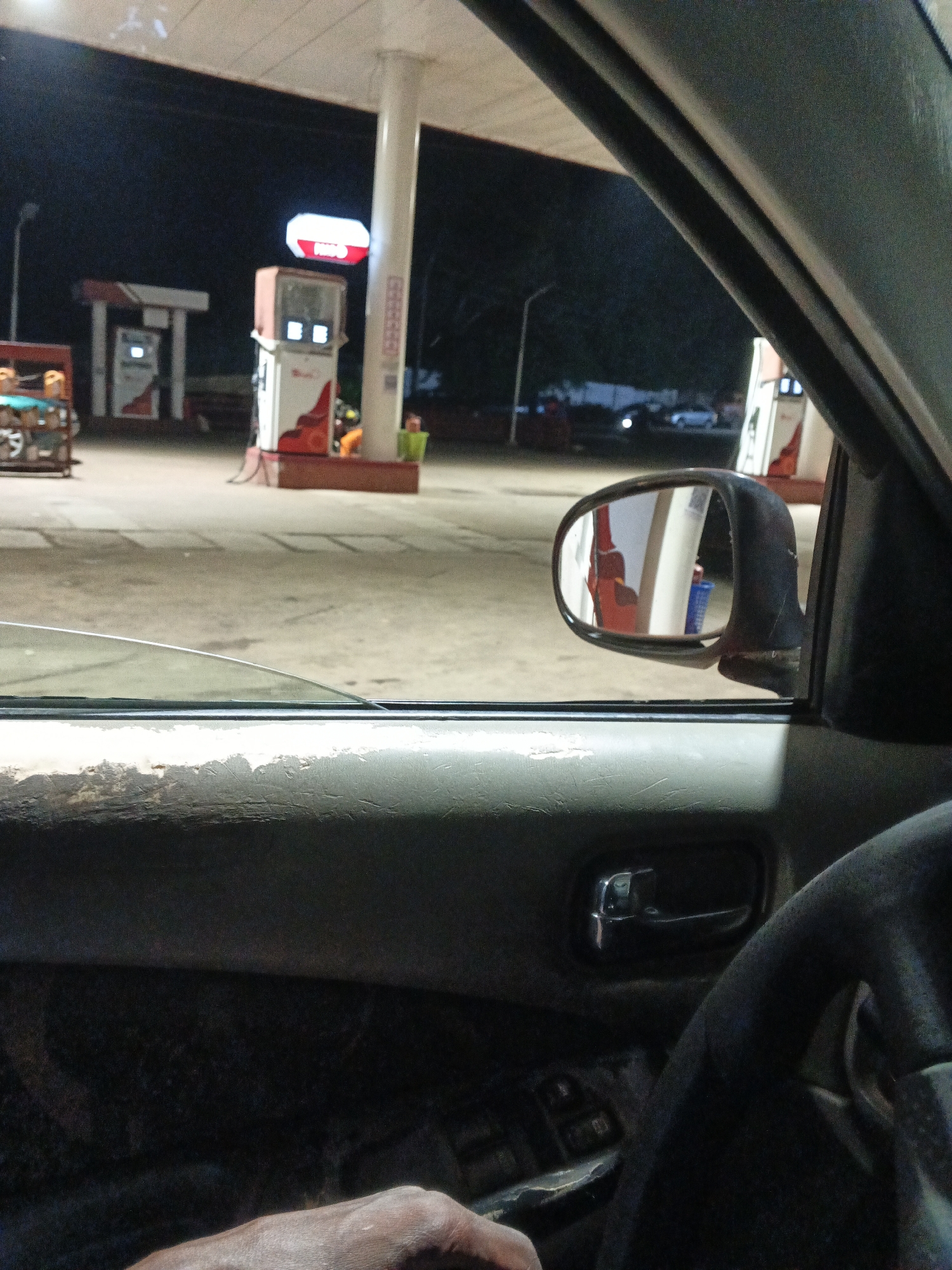 The image was captured from the interior of a car at night, likely at a gas station in Jos, Nigeria. The perspective is from the driver's seat, looking out through the passenger window and the side mirror.
In the foreground, the weathered texture of the car's interior door panel and a portion of the steering wheel are visible. A hand is resting on the door panel, suggesting the driver or a passenger.
Through the window and reflected in the side mirror, several gas pumps are illuminated, indicating the location is operational. The gas station appears to be of the common type found in Nigeria, with a covered canopy over the pumps. The ground is paved, and the overall lighting suggests it is night.
In the background, the surrounding environment is dark, with some faint lights and blurred shapes indicating other vehicles or structures. The side mirror reflects a gas pump and possibly a person near it, though details are obscured. The scene depicts a common nighttime scenario at a fuel station, possibly during a stop for refueling. The image does not contain any specific indicators of Jos, Nigeria, other than the provided context.