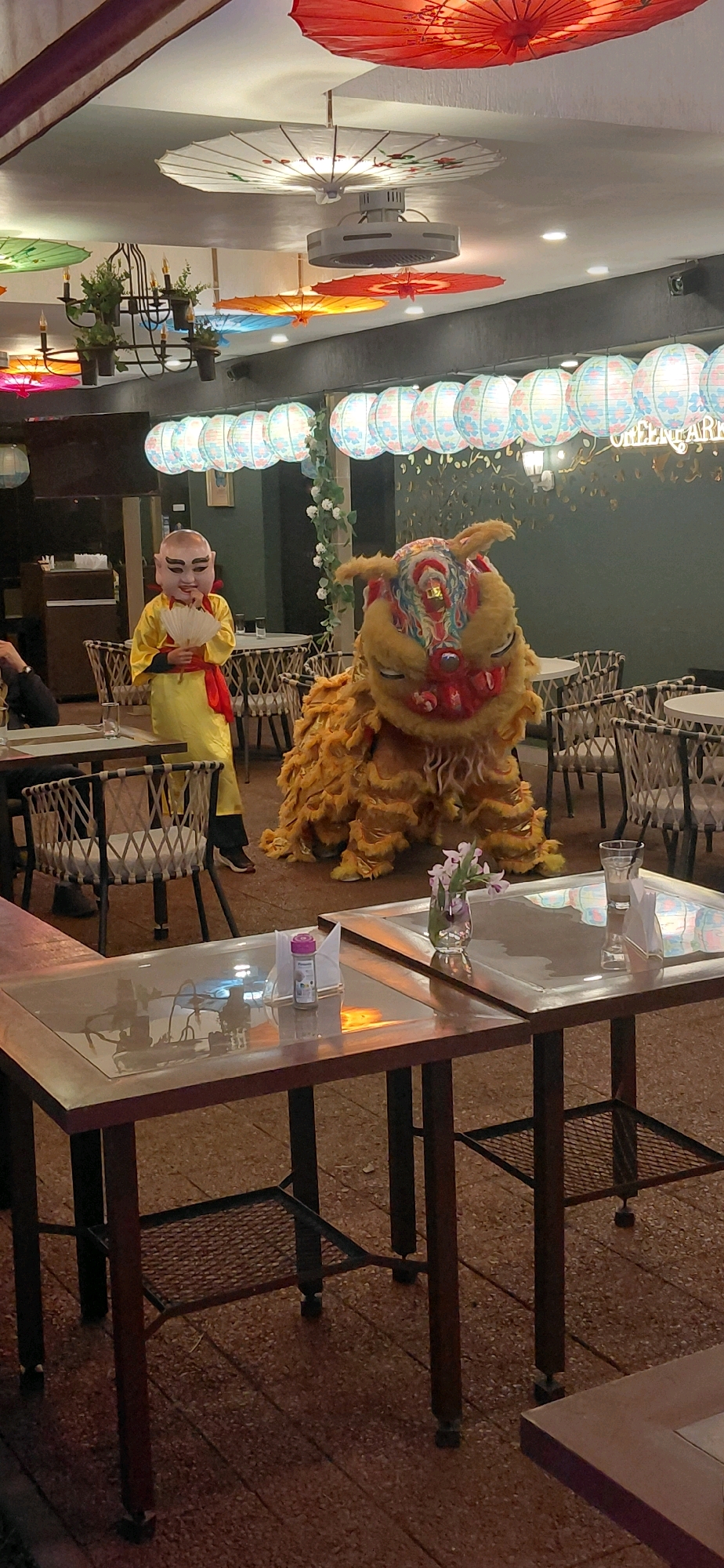 The image depicts the interior of a restaurant with an East Asian theme. In the foreground, two tables with dark brown wooden frames and glass tops are visible. One table has a small glass vase with pink flowers, a glass of water, and a napkin. The other table has a small spice container and a napkin. The floor is tiled with reddish-brown and grey stones.
In the mid-ground, a person in a yellow robe and a mask resembling a monk is holding a white fan. Next to them, a performer in a yellow lion costume is posed. The background shows a dimly lit restaurant with tables and chairs, decorated with numerous hanging paper lanterns and umbrellas in various colors, including red, white, blue, and green. A ceiling fan is visible above. The overall ambiance suggests a celebratory or cultural event. The question regarding the location in Encarnacion, Paraguay is outside the scope of image analysis.
