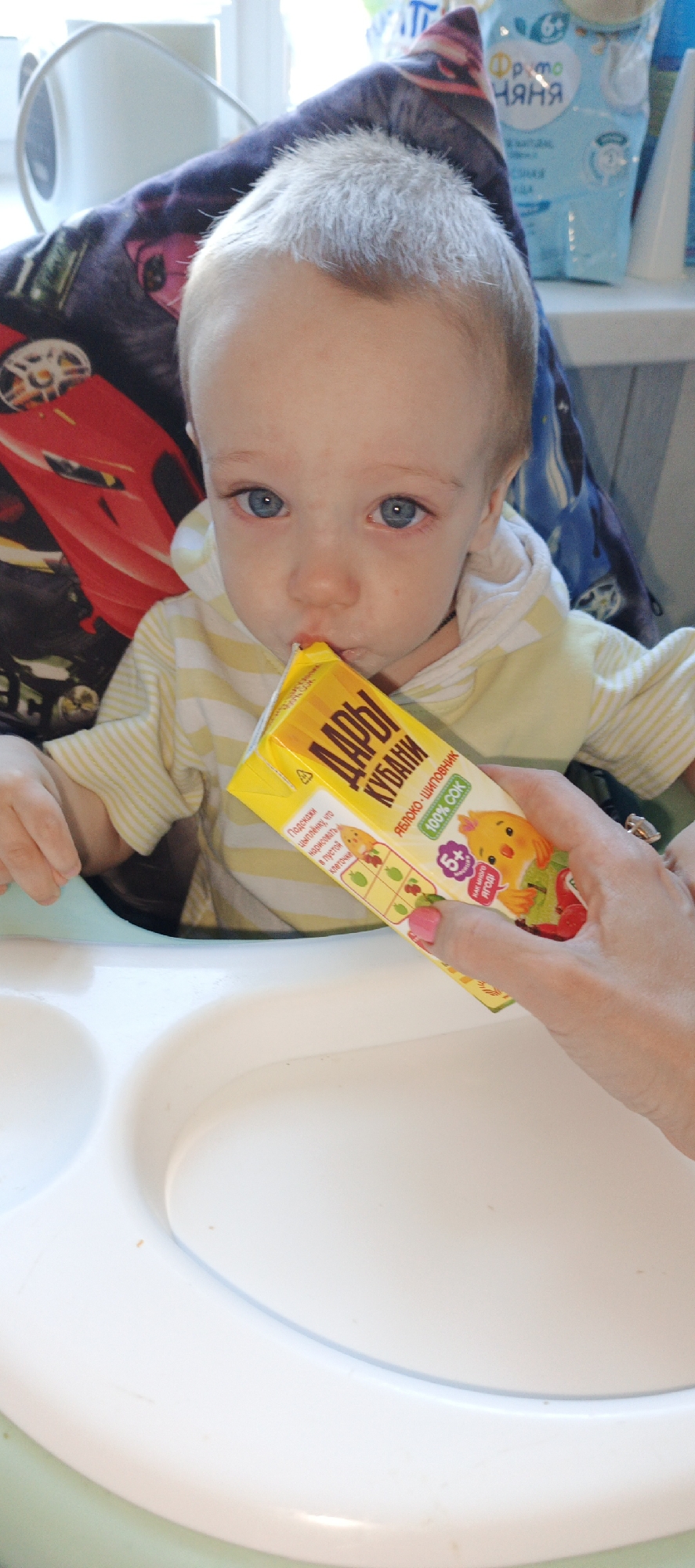 A young child with light hair and blue eyes is seated in a white high chair, actively drinking from a yellow juice box held by an adult's hand. The child is wearing a light yellow and white striped long-sleeve top. Their eyes appear somewhat red-rimmed.
The juice box is yellow with a cartoon chick and fruits, featuring Cyrillic text that reads "ДАРЫ КУБАНИ" (Gifts of Kuban), "ЯБЛОКО-ШИПОВНИК" (Apple-Rosehip), and "100% СОК" (100% Juice). Other text includes "6+" and "КАК МНОГО ЯГОД!" (How many berries!), along with a small interactive prompt: "Подскажи цыпленку, что нарисовать в пустой клеточке" (Tell the chick what to draw in the empty square). The adult's hand holding the juice box has pink nail polish.
The high chair has a wide, white tray with two divided sections in front of the child. Behind the child is a colorful patterned fabric, possibly a high chair cover or blanket, with red car designs. In the background, to the right, a light blue pouch or bottle is visible with Cyrillic text "Фруто Няня" (Fruto Nyanya), indicating a baby food or drink product. Bright natural light streams in from a window, suggesting it is daytime. The scene is set indoors, likely in a dining area or kitchen in Chaikovsky, Russia.
