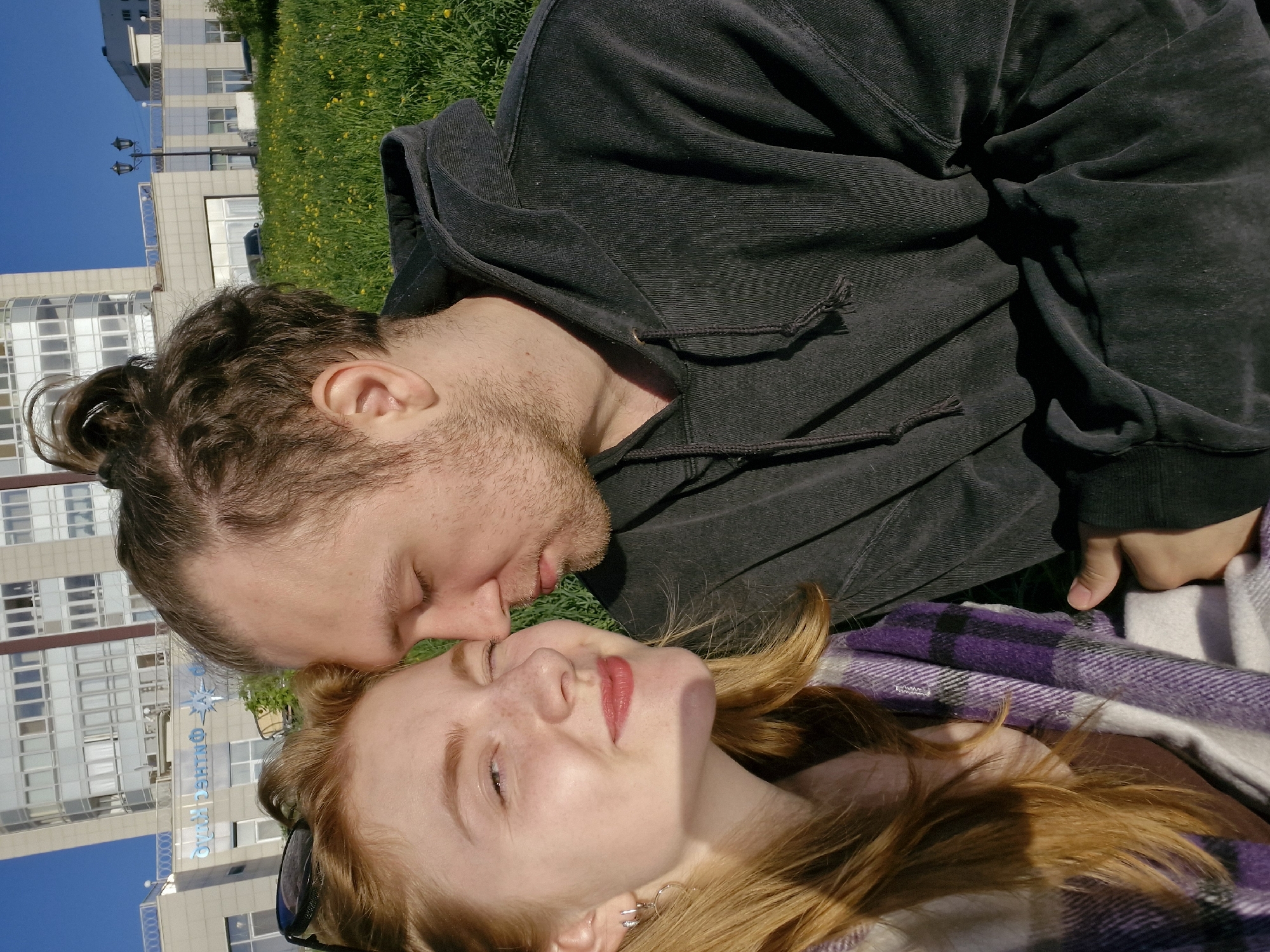 Two people, a male and a female, are lying on a green grassy area. The male is above, wearing a dark hoodie, with his face close to the female's face. The female is below, smiling with her eyes closed, lying on a purple and white plaid blanket. In the background is a building with multiple windows and a blue sky. This scene is in the city of Yekaterinburg, Russia, as indicated by the Russian writing on the building in the background.