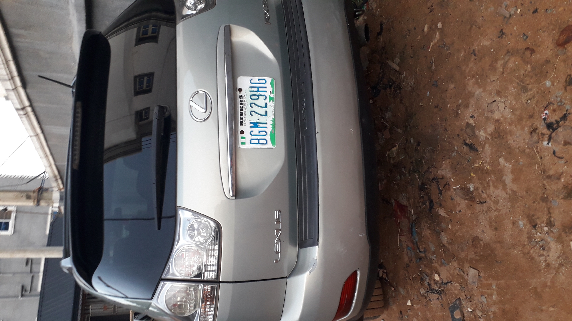 The image shows the rear of a silver Lexus SUV parked in what appears to be Nkpor, Nigeria, judging by the license plate which reads "BGM 229 HG" and identifies it as from Rivers State. The car is next to a building with a visible reflection in its windows. The ground is dirt and debris-covered. There are no people visible in the scene.
