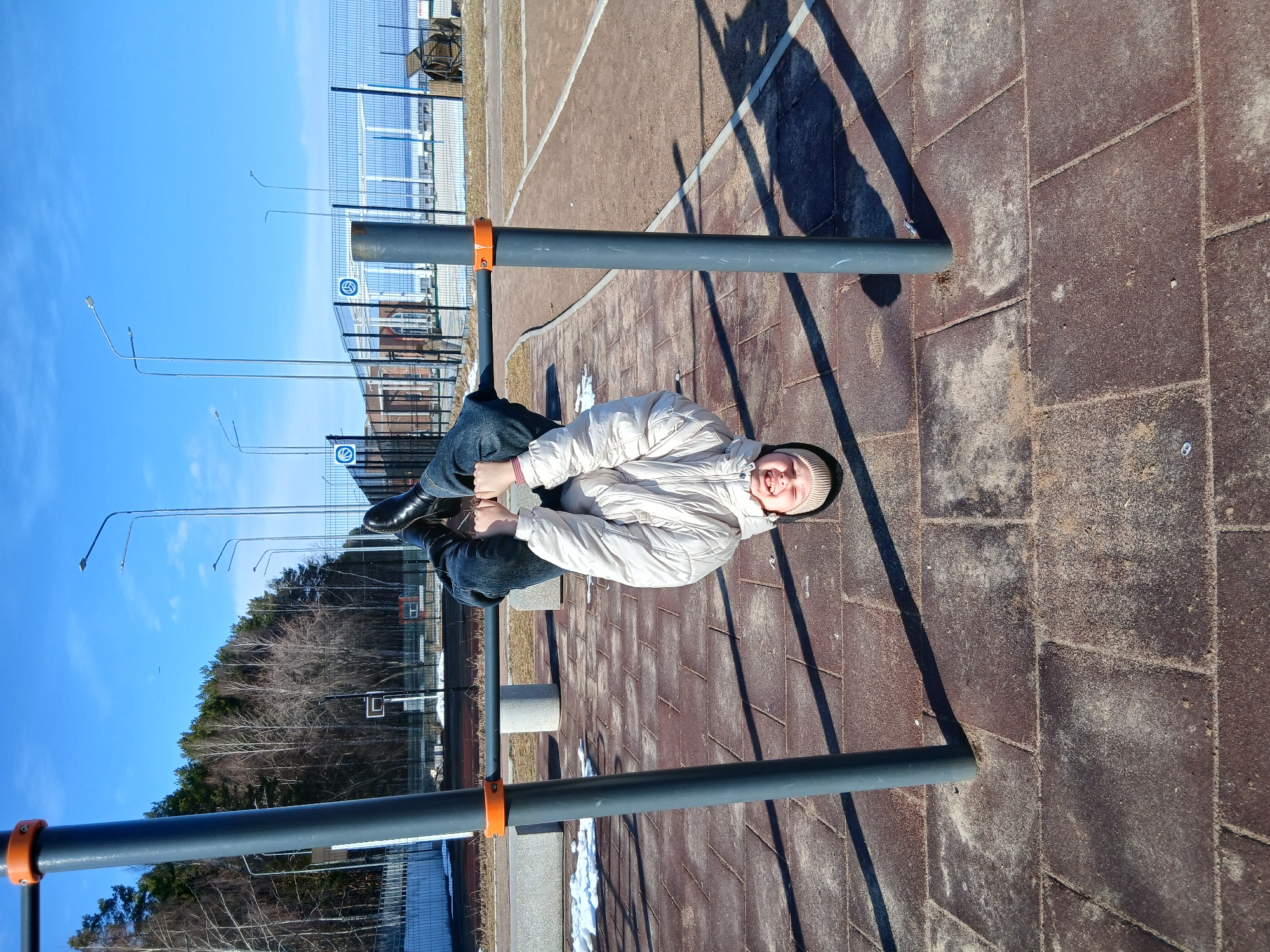 The image shows a person hanging upside down from a horizontal bar in an outdoor setting. The person is wearing a light-colored jacket, blue jeans, and dark shoes. They are smiling and looking at the camera. The bar is made of gray metal with orange accents at the connection points. The ground is paved with brown bricks. In the background, there is a metal fence, trees, and streetlights against a clear blue sky. The location is likely a public park or recreational area in the city.