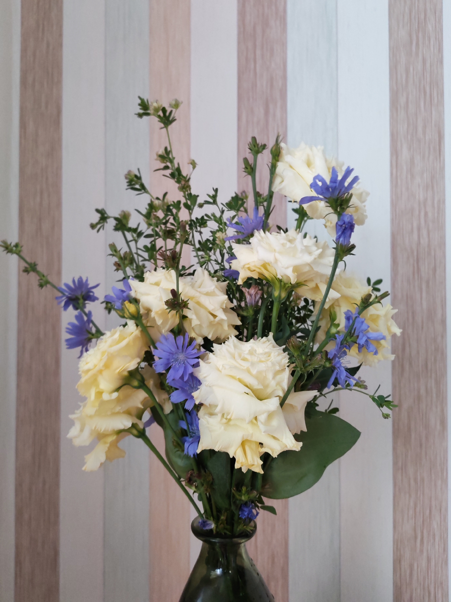 A bouquet of cream-colored roses and blue flowers sits in a dark green glass vase. The background is a striped wallpaper in shades of beige, pink, and white.