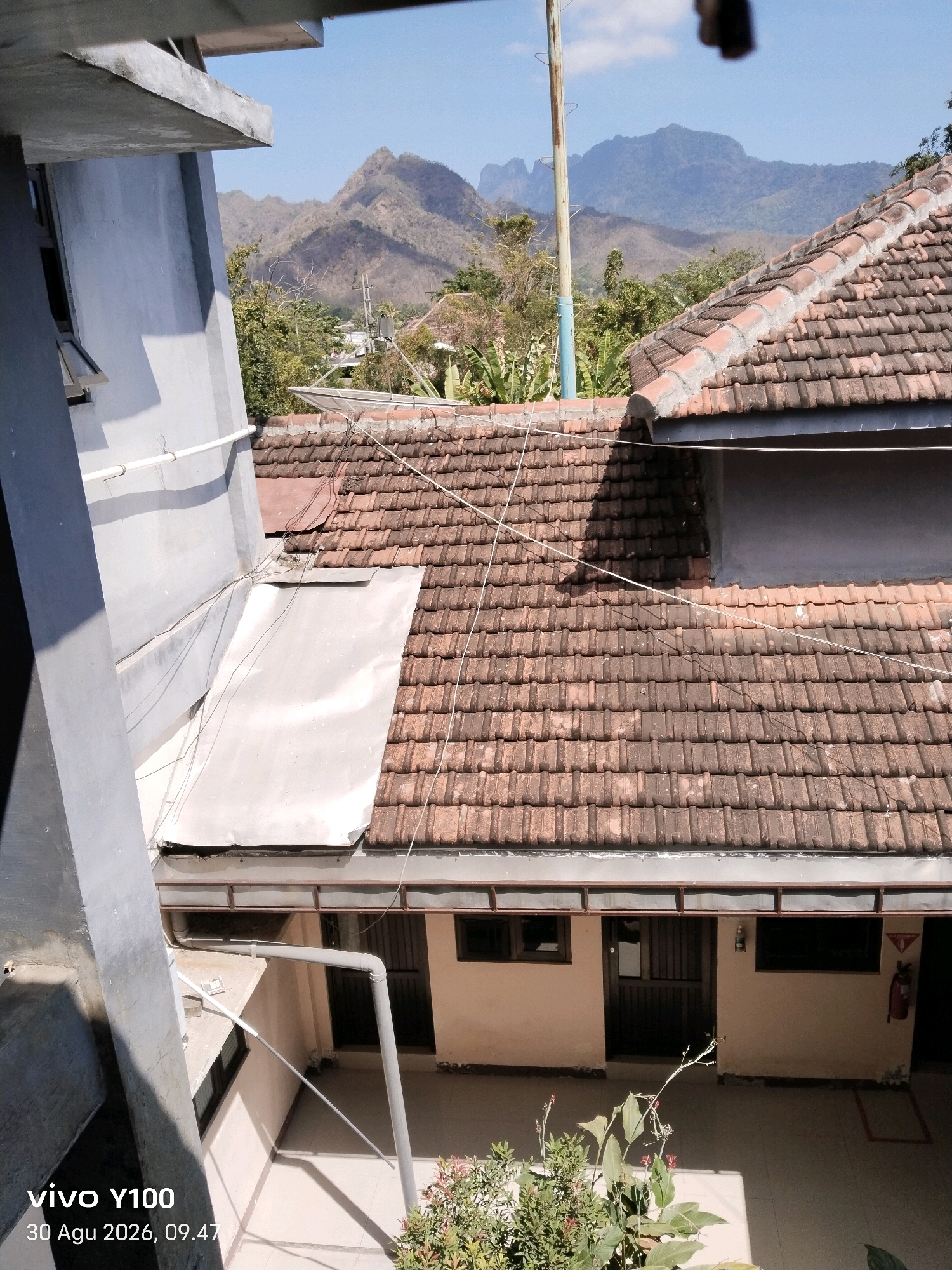 This is a daytime, eye-level shot from a high vantage point overlooking a tiled roof with a mountainous backdrop. The scene appears to be a residential area in Balung, Indonesia, with a clear blue sky and scattered clouds. The foreground shows a portion of a building's exterior, with a concrete structure on the left and a tiled roof on the right. The roof is made of weathered, reddish-brown clay tiles, with some sections appearing to have newer repairs or additions. Wires are strung across the roof, suggesting utility connections. Below the roof, a section of building facade is visible with windows and doors, leading into a courtyard area. A fire extinguisher is mounted on the wall in the lower right of the frame. In the distance, rolling hills and jagged mountain peaks dominate the horizon, covered in sparse vegetation. The overall impression is of a somewhat rural or semi-rural setting with a rustic architectural style.