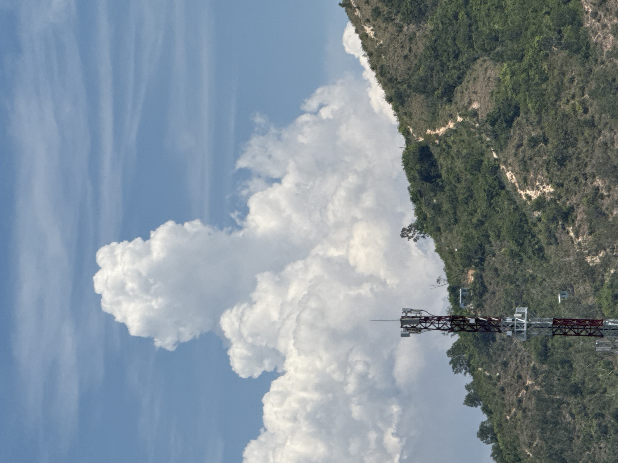 In Bac Nha Trang Ward, Vietnam, a red and silver cellular tower stands tall on a lush green hillside under a bright blue sky filled with dramatic white clouds. The tower, a lattice structure with multiple antenna arrays, is positioned on the lower left side of the frame, partially obscured by the foliage. A winding dirt path snakes up the side of the hill, indicating a hiking or access route. The hill is covered in a dense mix of trees and scrub, with patches of exposed earth. The sky above is a vivid blue, with a large, billowing cloud formation dominating the upper portion of the image.