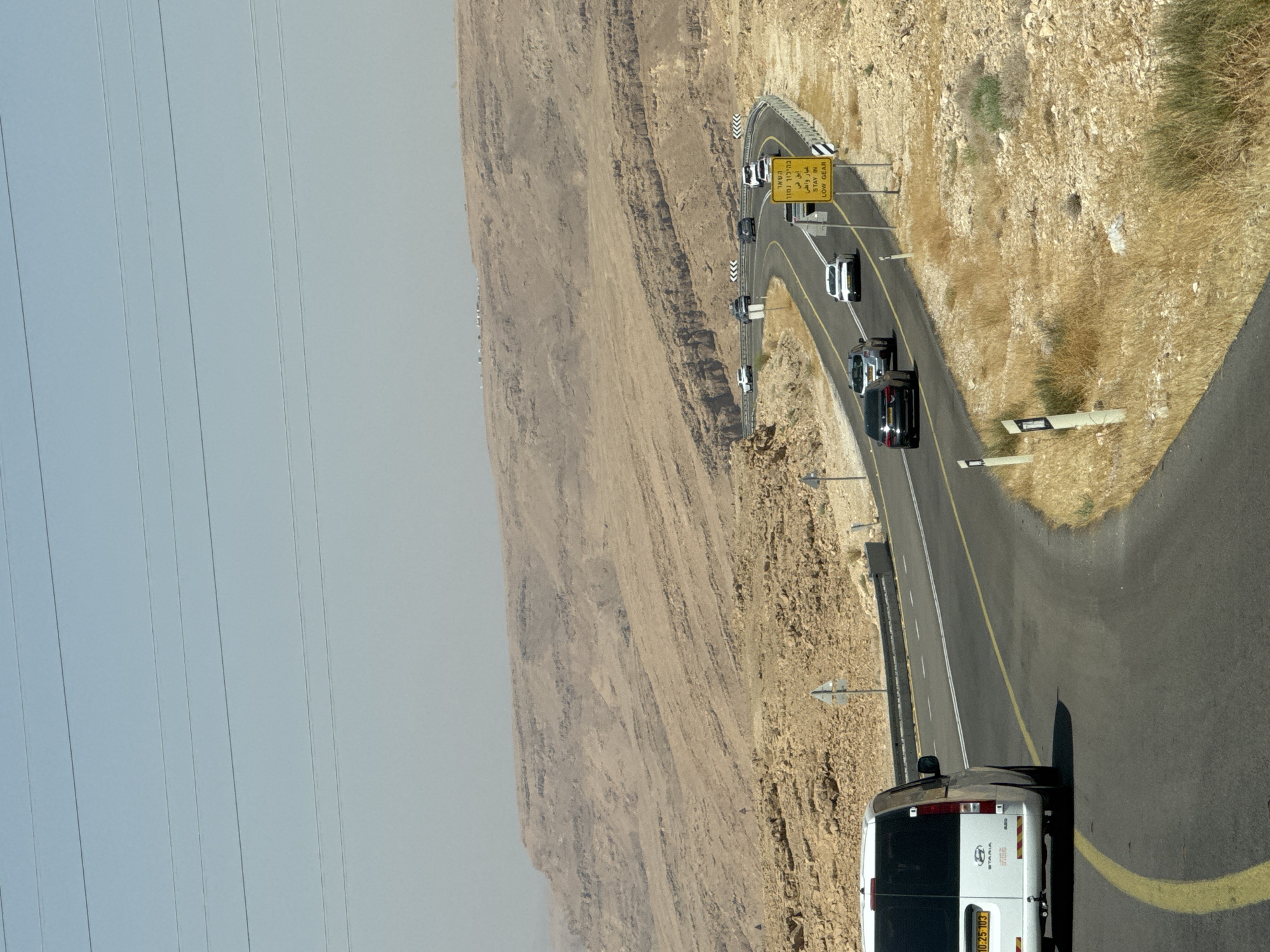 The image captures a winding road through a desert landscape, likely within the Ramat Negev Regional Council, Israel, given the Hebrew signage. A white van is prominently featured in the foreground, traveling on the right side of the road. Several other vehicles, including cars and SUVs, are visible on the asphalt road ahead, navigating the curves. The road is bordered by guardrails and accompanied by a yellow advisory sign written in Hebrew and English, stating "STAY IN LOW GEAR," indicating a steep incline or descent. The environment is arid, characterized by dry, rocky terrain and sparse vegetation, with layered, eroded rock formations forming the backdrop under a clear, hazy sky. Overhead power lines are visible stretching across the upper portion of the frame. The overall scene depicts a travel route through a rugged, desert environment.