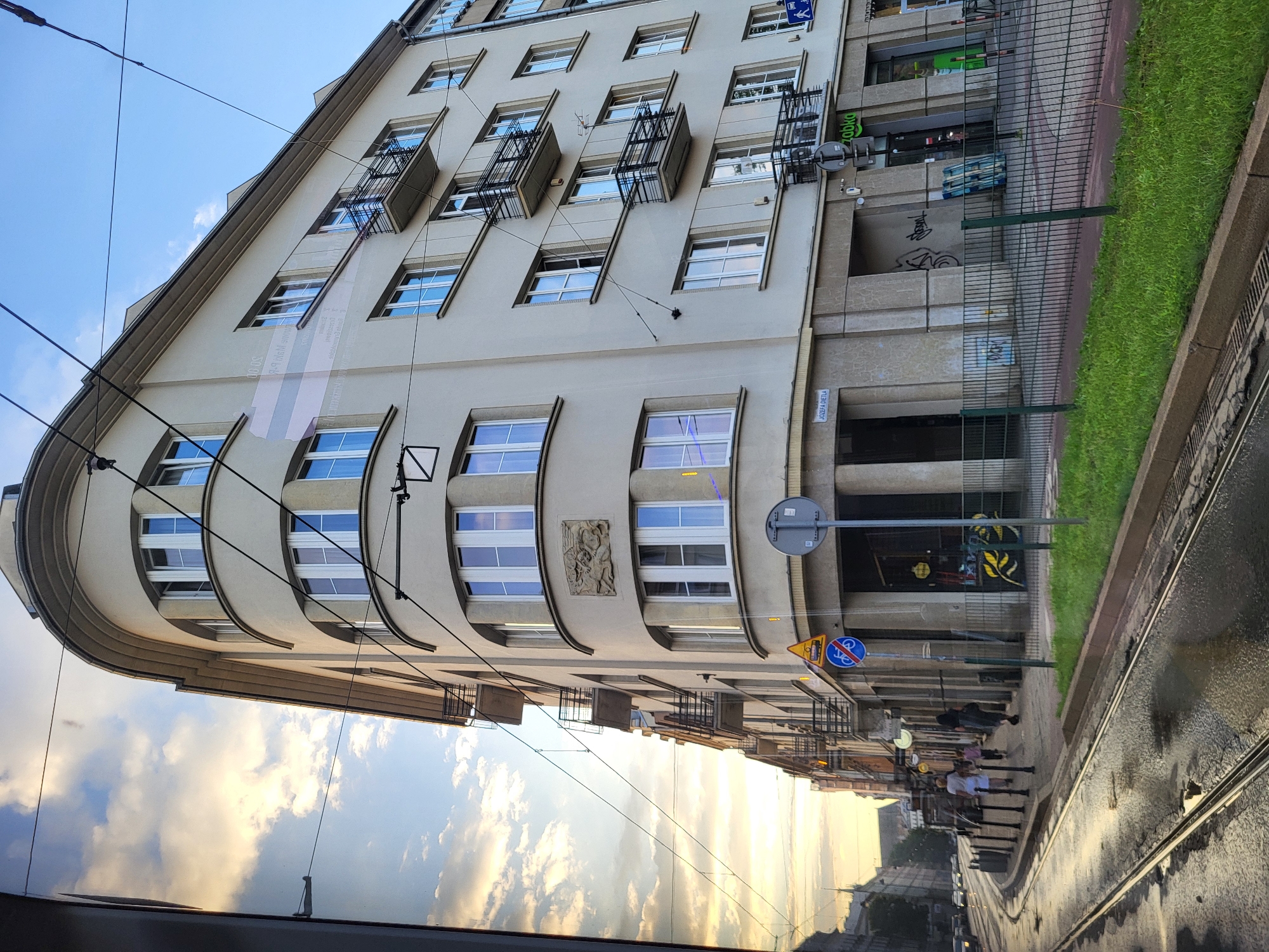 The image shows a curved building on a city street with tram tracks in the foreground. The building is light-colored with multiple floors and windows, and has several balconies. There are overhead wires for the tram system crisscrossing the sky. The street appears wet, reflecting the sky and buildings. Several people are walking on the sidewalk in front of the building. The sky is partly cloudy with warm, golden light suggesting it is either sunrise or sunset. There are signs on the building, including one that reads "JOZEFA DIETLA" and another with the logo for "ZABKA." The overall scene depicts a street view in an urban environment.