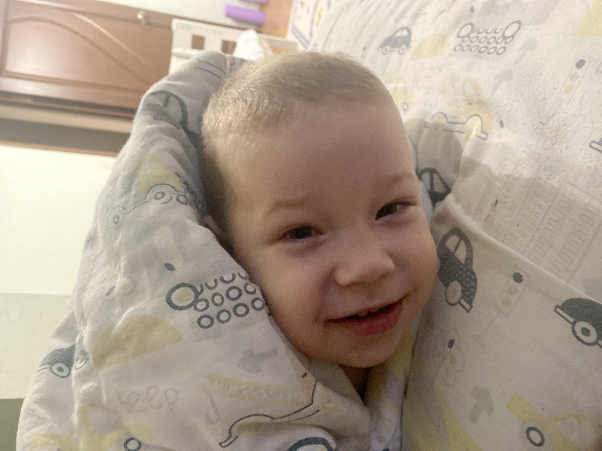 A young child with fair skin and blonde hair is wrapped in a patterned blanket. The blanket features images of cars, circles, and text. The child is smiling slightly, with their eyes partially closed. The background includes a wooden cabinet. The exact location within Sochi, Russia, cannot be determined from this image.