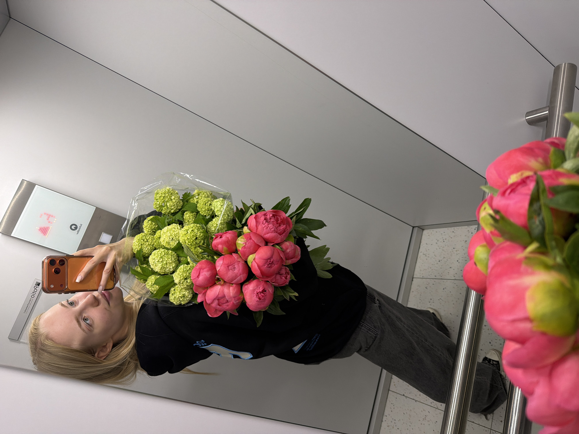 A reflection in a mirror shows a woman in Unknown, Unknown. She has long blonde hair and is holding a bouquet of pink and green flowers. She is wearing a black sweatshirt and gray jeans. The mirror reflects a modern interior with white walls and stainless steel accents. The woman is holding a smartphone, presumably taking the photograph.