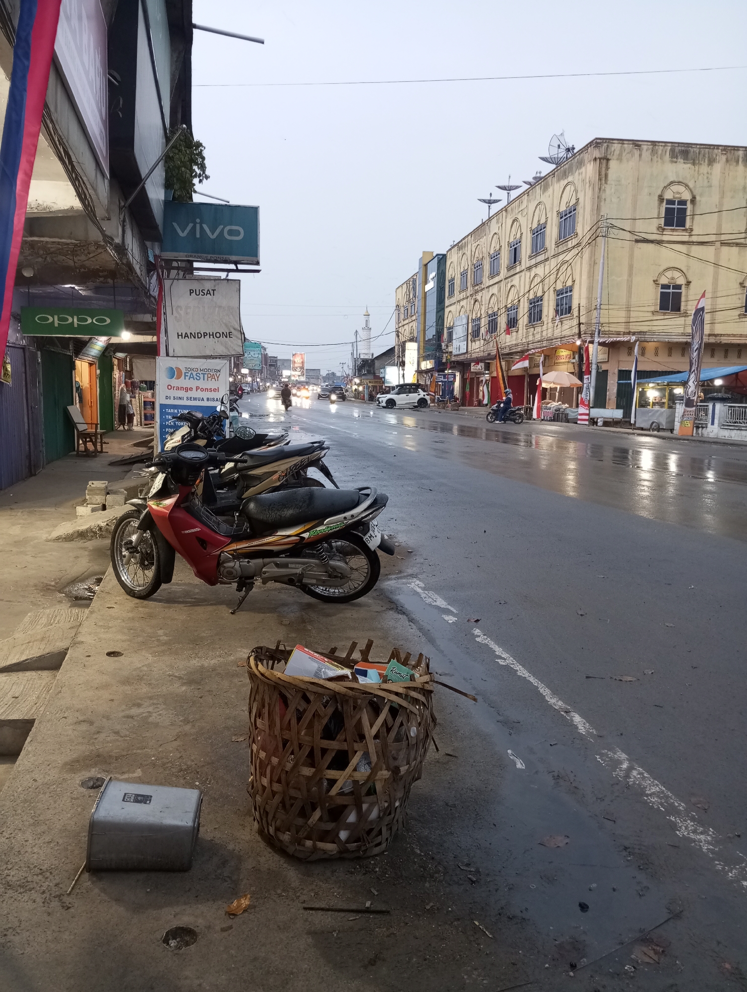 The image captures a street scene in Air Molek, Indonesia, during what appears to be overcast or early morning light, with the ground wet from rain. In the foreground, two motorcycles are parked on a concrete sidewalk. The motorcycle closest to the viewer is red and silver, with the Honda logo visible. Beside it is a large, woven basket filled with various items, including boxes and plastic containers. To the left of the basket, a silver metal box lies on the ground.
The sidewalk is adjacent to a street where traffic is moving. Several vehicles, including cars and motorcycles, are visible on the wet asphalt. Buildings line the street. On the left side of the road, there are shops with signage, including an "OPPO" logo and a "VIVO" sign. Further down the street, a multi-story building with arched windows and satellite dishes on its roof is prominent. Various flags, some appearing to be Indonesian national flags, are displayed in front of some establishments. The overall impression is of a typical commercial street in a Southeast Asian town.