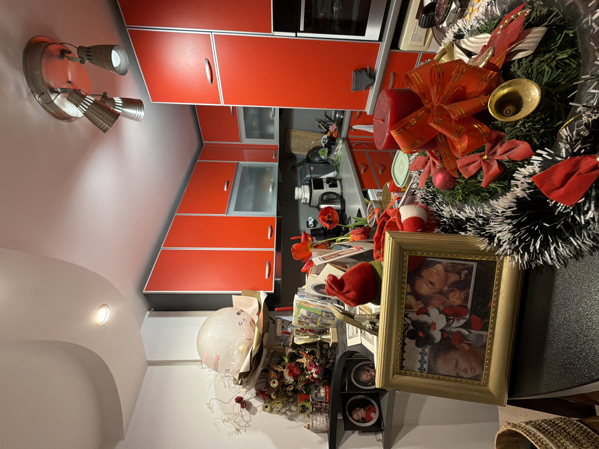The image shows an interior scene. A kitchen with red cabinets is visible in the background. In the foreground, a cluttered surface holds various objects including a picture frame, a balloon, flowers, and a Christmas wreath. The location is not identifiable as being in Varna, Bulgaria.