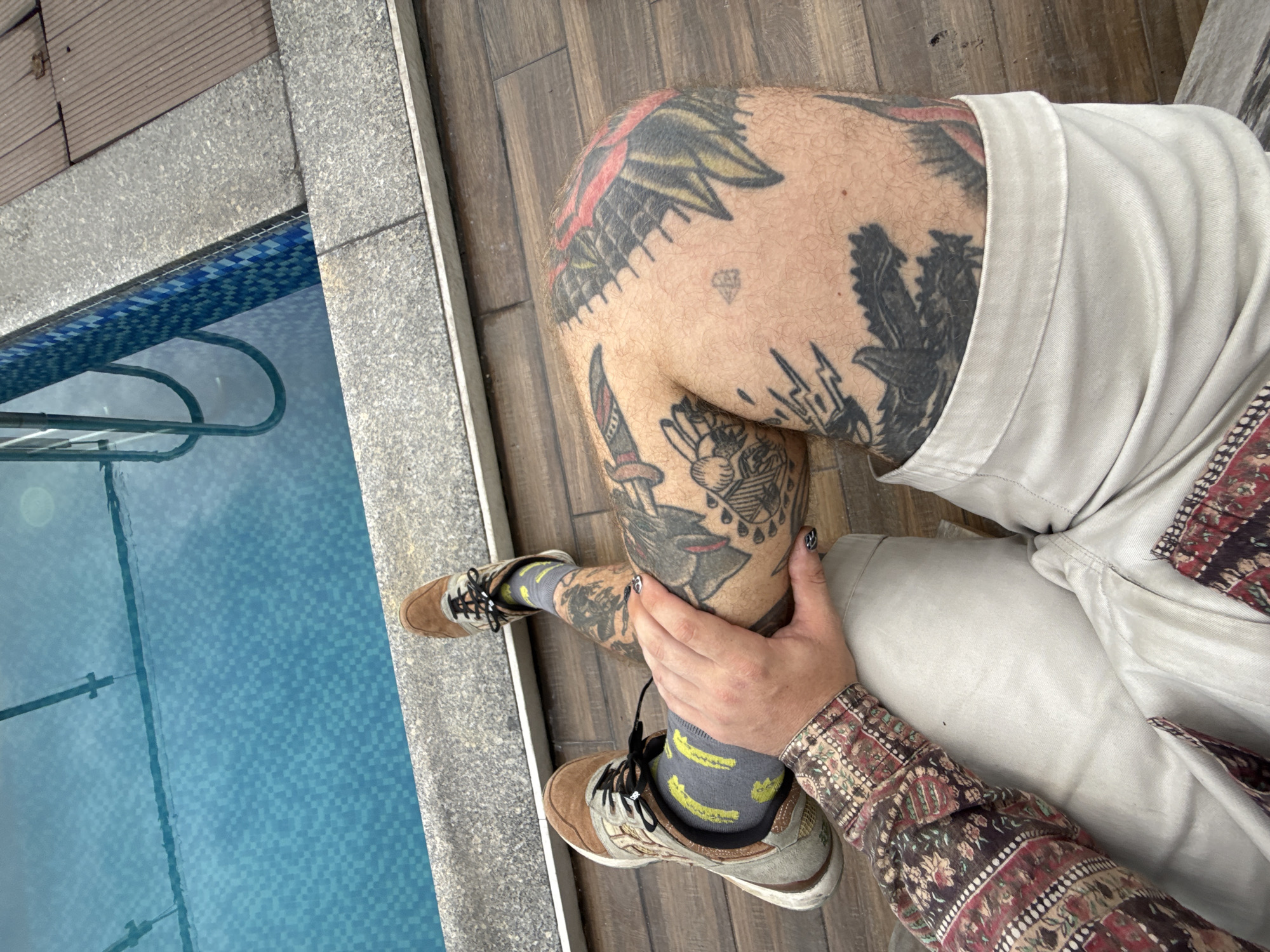 A person sits at the edge of a pool. The person has numerous tattoos on their leg and wears beige shorts, a patterned shirt, socks with banana patterns, and tan and gray sneakers. They are holding their knee. The pool is tiled with blue and white mosaic tiles. The ground surrounding the pool is a mix of wood and gray concrete. It is daytime. Although the location is allegedly in Da Nang, Vietnam, there is no evidence that corroborates that assertion.