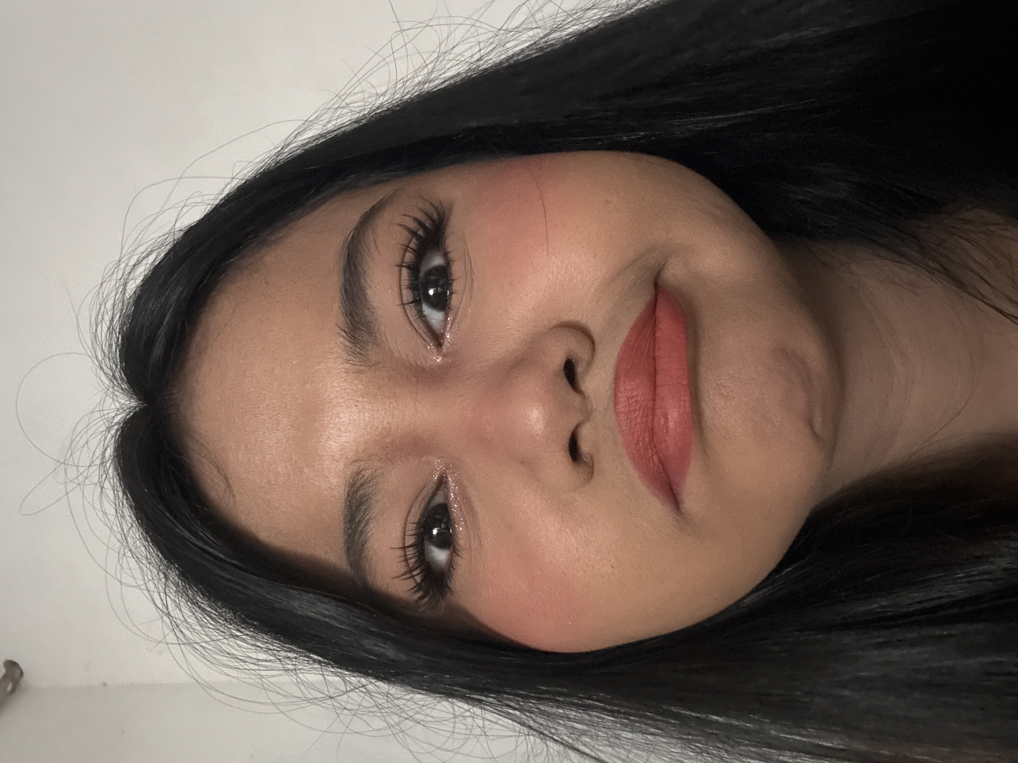 A close-up, frontal, waist-up shot shows a young Indonesian woman with long, dark hair and fair skin. She is wearing heavy eye makeup, including false eyelashes, and coral-toned lipstick. Her cheeks are heavily rouged with blush. Her expression is one of slight pout or displeasure, with her mouth downturned. Her eyes are wide open and looking directly at the camera. The background is a plain, white wall. The image was taken in North Jakarta, Indonesia.