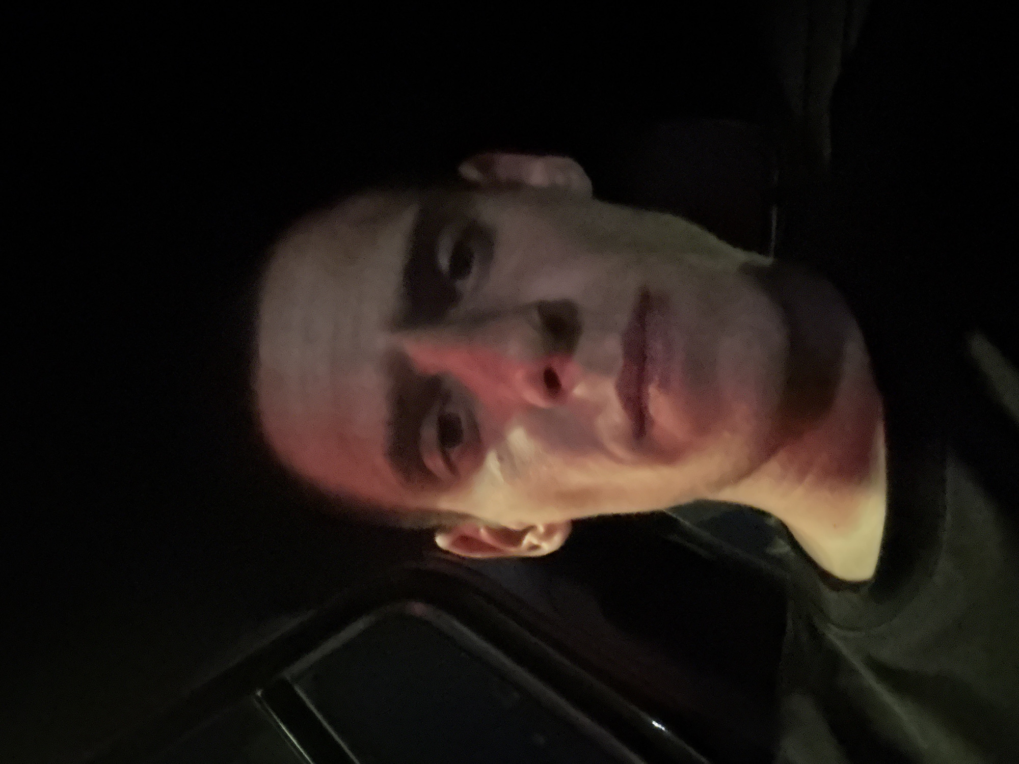 The image is a close-up, self-taken photograph of a male subject's face, appearing to be taken inside a vehicle at night. The subject has short, dark hair, dark eyes, and is wearing a black t-shirt. A red light is reflecting on the left side of his face, illuminating his cheek and nose. The background is dark, with the interior of a car visible on the left side of the frame, suggesting the subject is in the driver's seat. There is no identifiable location in Sofia, Bulgaria, within the image.