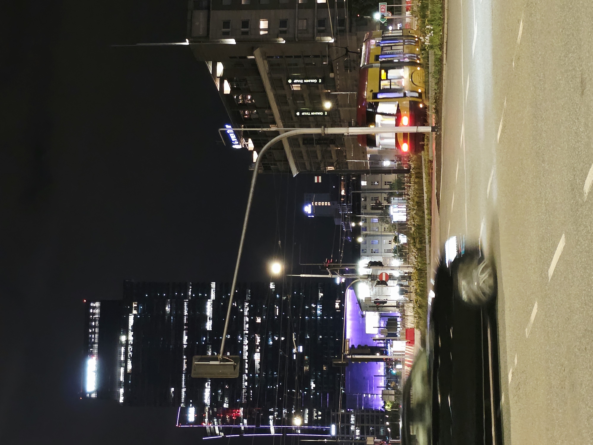 The image depicts a night scene in Warsaw, Poland, likely in the city center. Dominating the background is a tall, modern skyscraper with numerous illuminated windows, suggesting an office building. In the middle ground, a yellow and red tram is stopped at a crossing, its headlights and interior lights visible. To the right of the tram, a multi-story building displays the "GOLDEN TULIP" hotel signage. A black car is in motion in the foreground, blurred due to its speed, suggesting it is driving on a city street at night. The street is paved, with clear lane markings. Traffic lights and street lamps are visible, contributing to the urban nighttime atmosphere. The sky is dark, indicating it is late evening or night. The scene appears to be an active urban intersection with significant infrastructure and transportation.