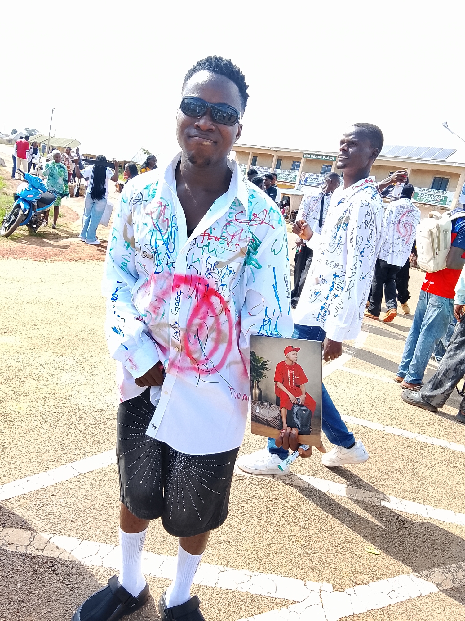 A man in a white, graffiti-covered shirt and black shorts stands in the foreground, smiling at the camera. He is wearing sunglasses and black slip-on shoes with white socks. In his right hand, he holds a framed picture of a young man in a red outfit. Behind him, other people are visible in a street setting, with buildings and a blue motorcycle in the background. The ground is paved with asphalt and marked with white lines. The scene appears to be outdoors on a sunny day. The location is identified as Tambes Mile Four, Nigeria.