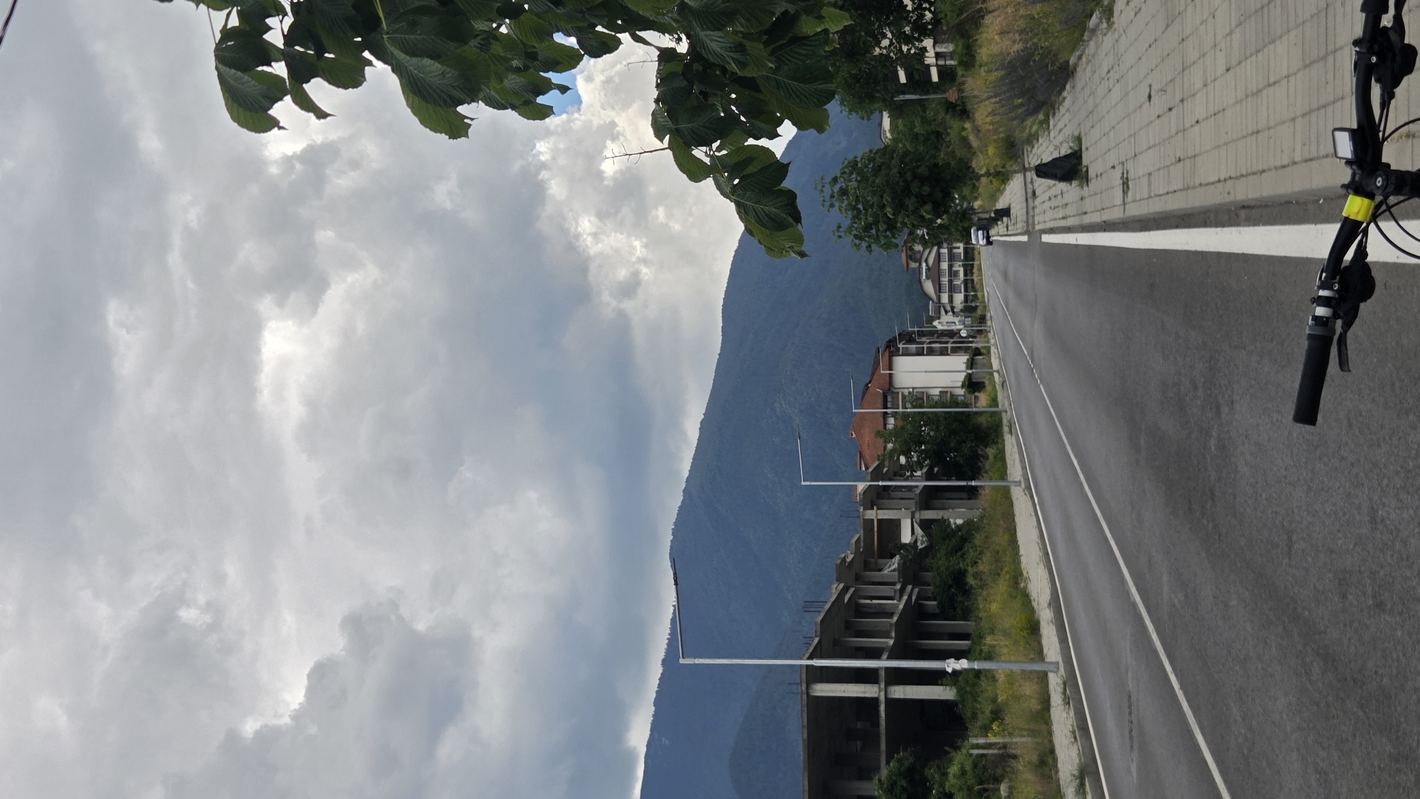 A vertical, eye-level, medium shot captures a street in Bansko, Bulgaria, under a cloudy sky. The road stretches into the distance, bordered by a paved sidewalk on the right and a strip of dry grass and low vegetation on the left. Partially constructed buildings, with exposed concrete pillars and floors, are visible on the left side of the road. On the right, modern apartment buildings with white facades and dark roofs line the street. In the background, a large, tree-covered mountain dominates the horizon. The foreground shows the handlebars of a bicycle, with a yellow zip tie on the left grip. The scene appears to be captured on an overcast day.
