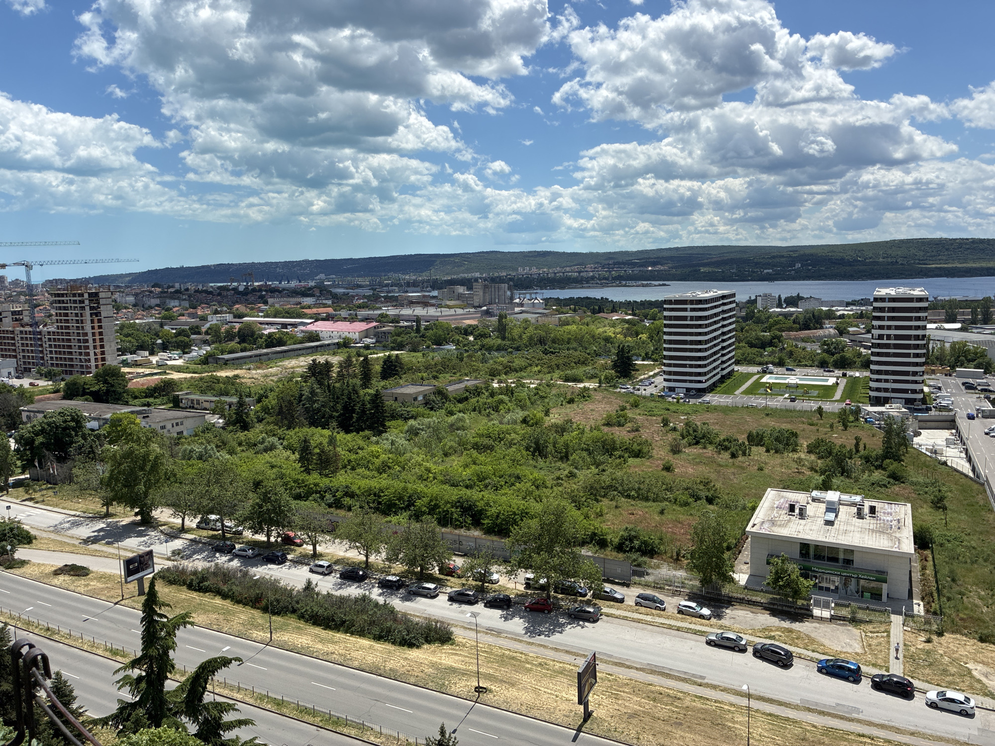 The image depicts an elevated view in Varna, Bulgaria, characterized by a mix of urban development and natural elements under a sky with scattered clouds.
In the foreground, a multi-lane road is visible, lined with trees and several parked cars. There are also two signboards.
Beyond the road is a green space with scattered buildings, including two tall, white apartment complexes with horizontal black bands. Closer is a lower-rise building with a sign in Bulgarian, "Медицински център Хейлан". A person walks along the path.
In the middle distance, a city skyline extends towards the horizon, and a body of water is visible beyond the city, appearing to be the Black Sea. A crane is present in the top left corner of the image.