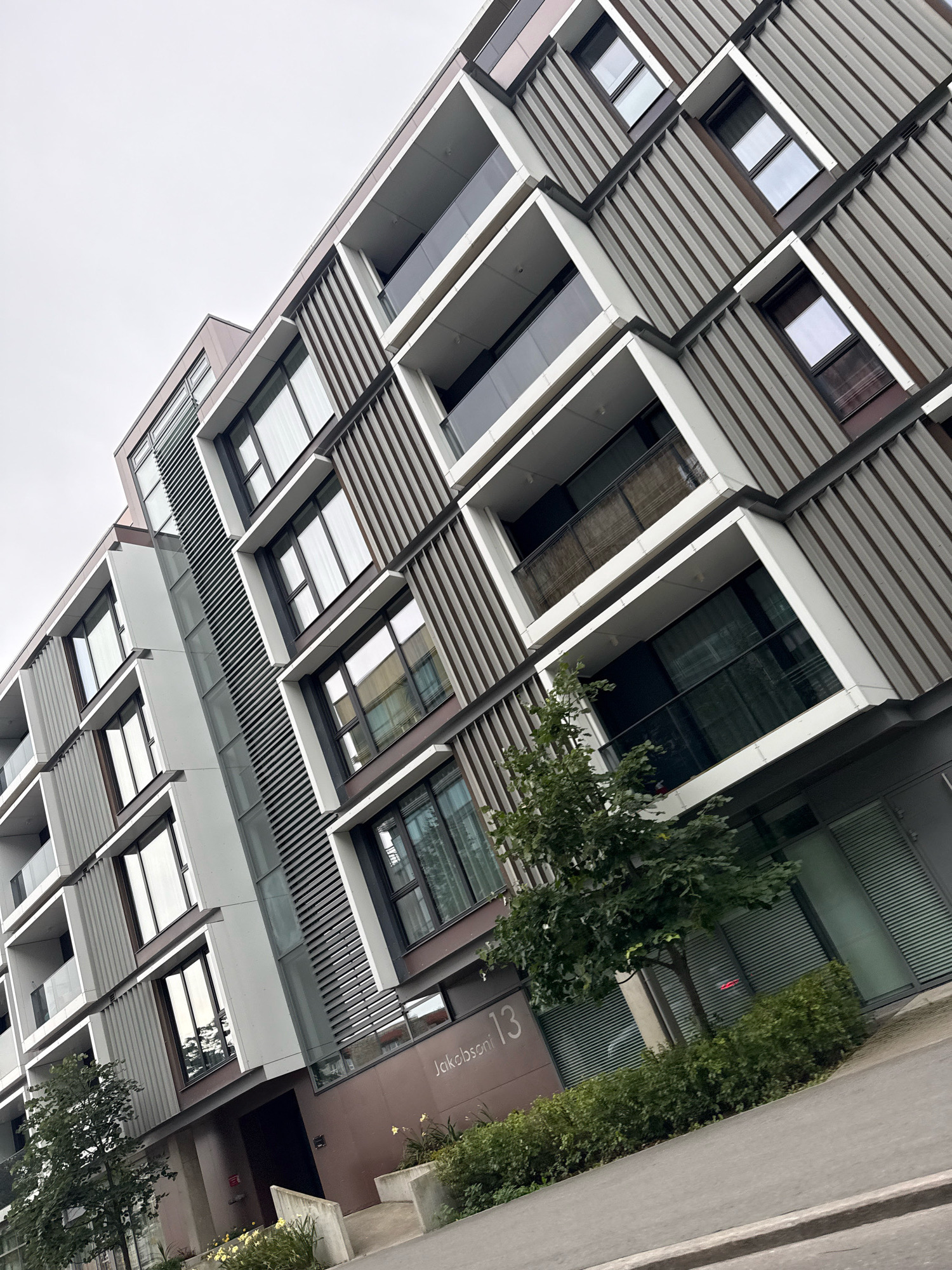 A low-angle, full shot captures the modern, multi-story apartment building at Jakobsoni 13 in Tallinn, Estonia. The building's facade is characterized by a combination of metallic grey corrugated panels, white framed balconies with glass railings, and dark-framed windows. The architecture features a staggered design with protruding and recessed sections. A young tree with green foliage stands to the left of the building, with a neatly trimmed hedge and some low-lying yellow flowers in planters in front of the entrance. The ground in front of the building is paved, with a sidewalk and a road visible. The sky is overcast and grey. No people are visible in the frame.